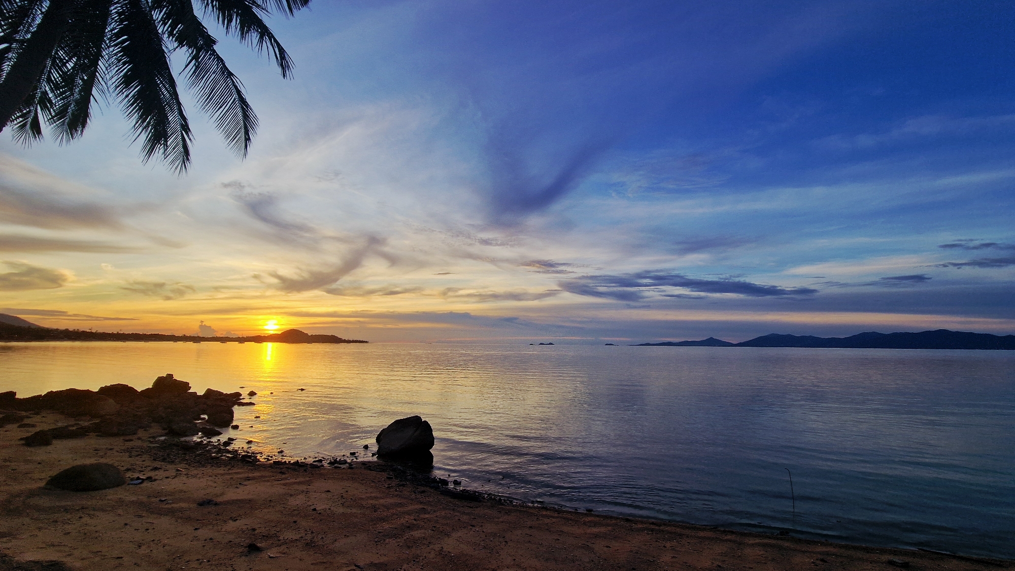 This is a wide-angle, eye-level, horizontal photograph taken at dusk in Baan Bang Rak, Thailand. The scene depicts a serene beach with a calm ocean and a vibrant sky. In the foreground, a sandy beach is visible with several dark rocks scattered along the shoreline. To the left, a portion of a silhouetted palm tree hangs over the scene, its fronds reaching down towards the water.
The ocean occupies the middle ground, reflecting the warm hues of the setting sun. The sun itself is low on the horizon, positioned over a distant landmass that appears to be an island or the mainland. The water is calm with gentle ripples. Several small islands or landforms are scattered in the distance across the ocean.
The sky is a dominant feature, transitioning from warm orange and yellow tones near the horizon where the sun is setting, to shades of blue and purple as it ascends. Wispy clouds, tinged with the colors of sunset, stretch across the sky, adding depth and texture. The overall impression is one of tranquility and natural beauty at the end of the day.