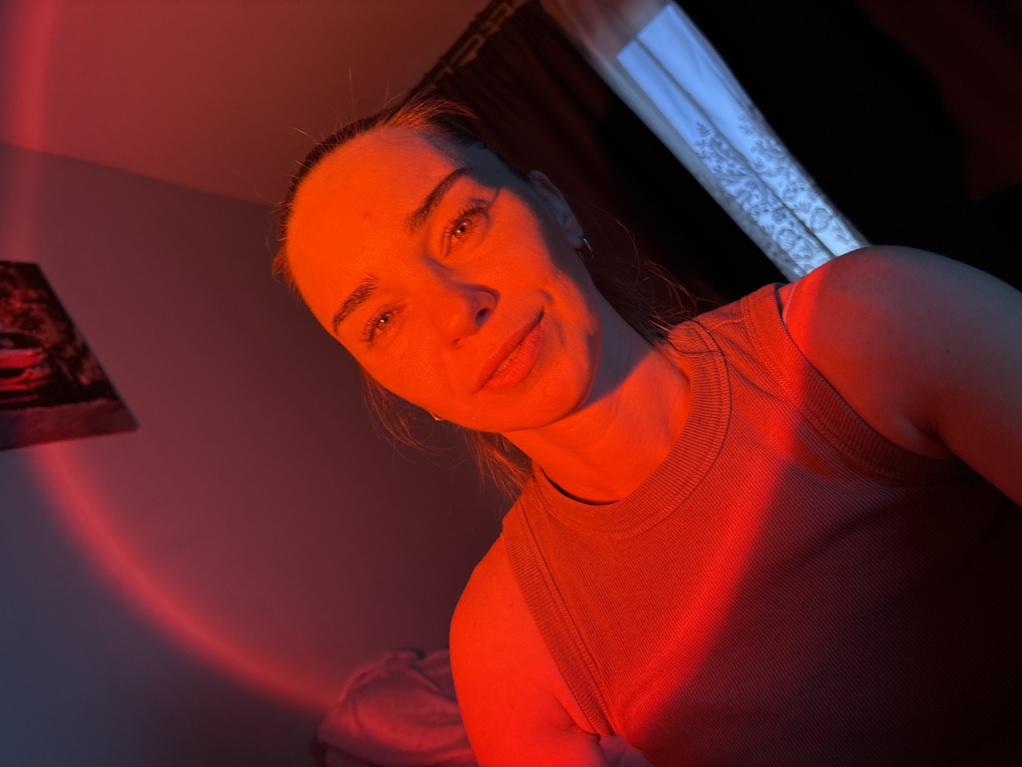 A selfie-style, close-up portrait of a woman is captured in a dimly lit room with a prominent red light casting a strong glow on her face and upper body. The woman, with her hair pulled back, gazes towards the camera with a slight smile. She is wearing a ribbed, tank-top style shirt. The background shows a wall with a framed image or artwork at the top left, illuminated by the same red light, and what appears to be a window with a curtain to the right. The overall ambiance is moody and atmospheric due to the dramatic lighting. The location is not specified in the image or its metadata.
