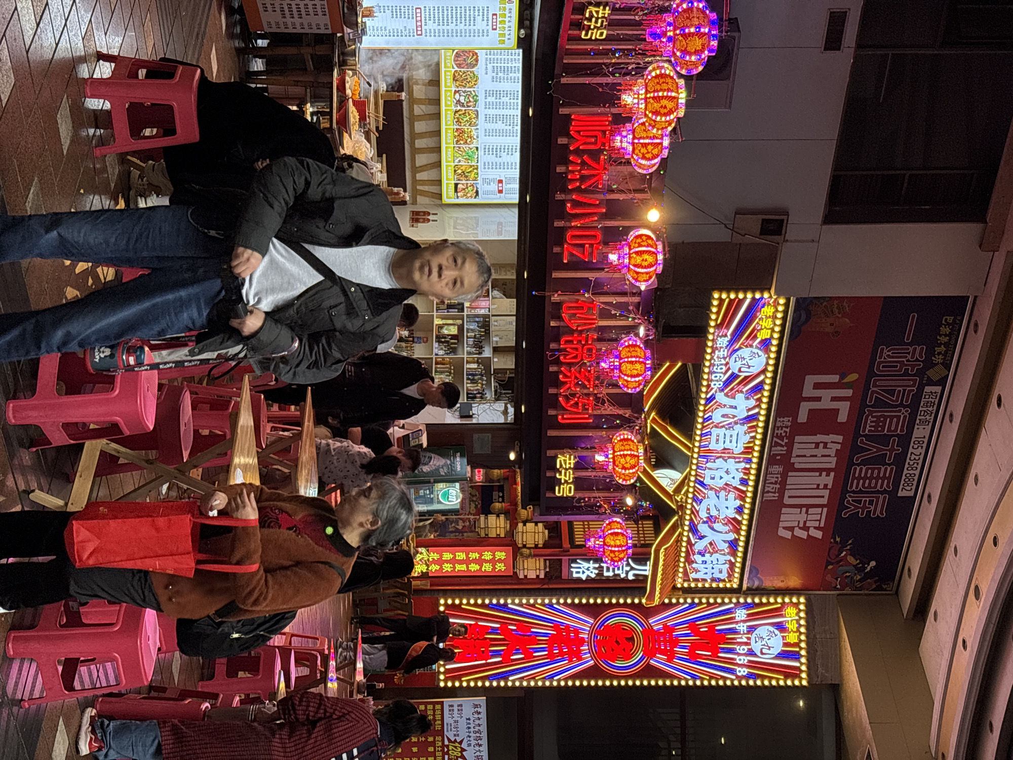 The image shows a vibrant street scene at night in the Yuzhong District of Chongqing, China, characterized by numerous bright neon signs in Chinese characters and colorful hanging lanterns. Several people are present, including a man in a dark jacket and blue jeans looking at the camera, and an elderly woman in a brown jacket carrying a red bag. Both are seated on small red plastic stools, which are widely distributed across the scene. A menu board is visible, indicating a food establishment. Other people can be seen dining and walking around, suggesting a bustling commercial environment.
