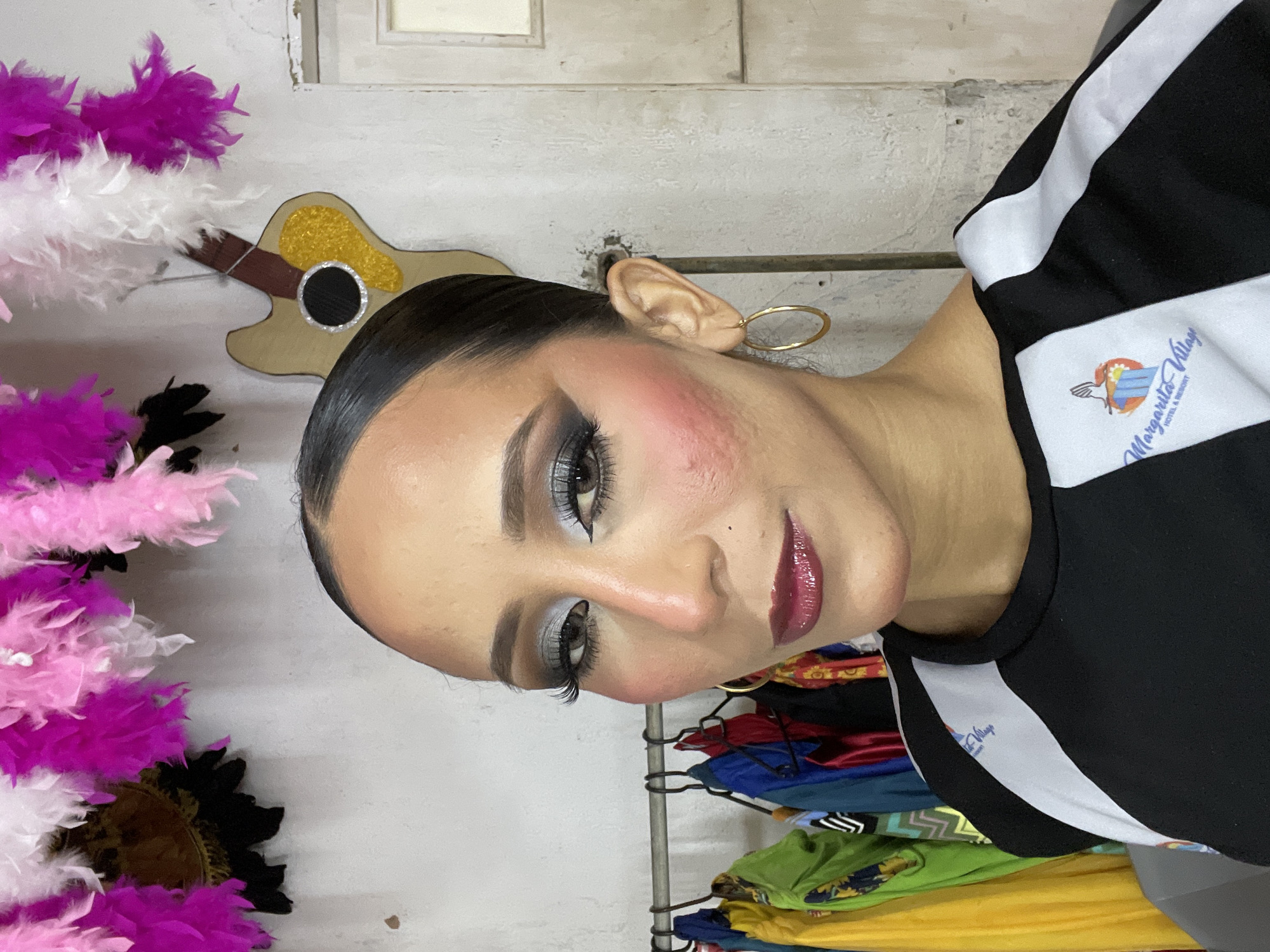 The image shows a close-up of a woman with elaborate makeup, including dramatic eye makeup and glossy lipstick. She is wearing a black and white top with a logo that reads "Margarita Village Hotel & Resort." Behind her, there is a rack of colorful garments, a decorative guitar, and a wall adorned with pink and white feathers. The scene appears to be set in a studio or dressing room, possibly in Porlamar, Venezuela, given the presence of the "Margarita Village Hotel & Resort" logo. The lighting is bright, highlighting the woman's features and the details of her attire and surroundings.