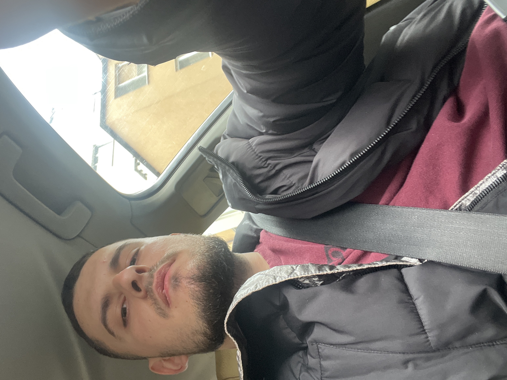 A selfie of a young man with short brown hair and a beard is taken from inside a car. He is wearing a maroon shirt, a black puffy jacket, and a seatbelt. The car's sunroof is open, revealing a building exterior. The photo's setting is likely a vehicle in an urban environment, although a more precise location cannot be determined from the image alone. There is nothing in the image to suggest this is in Burgas, Bulgaria.