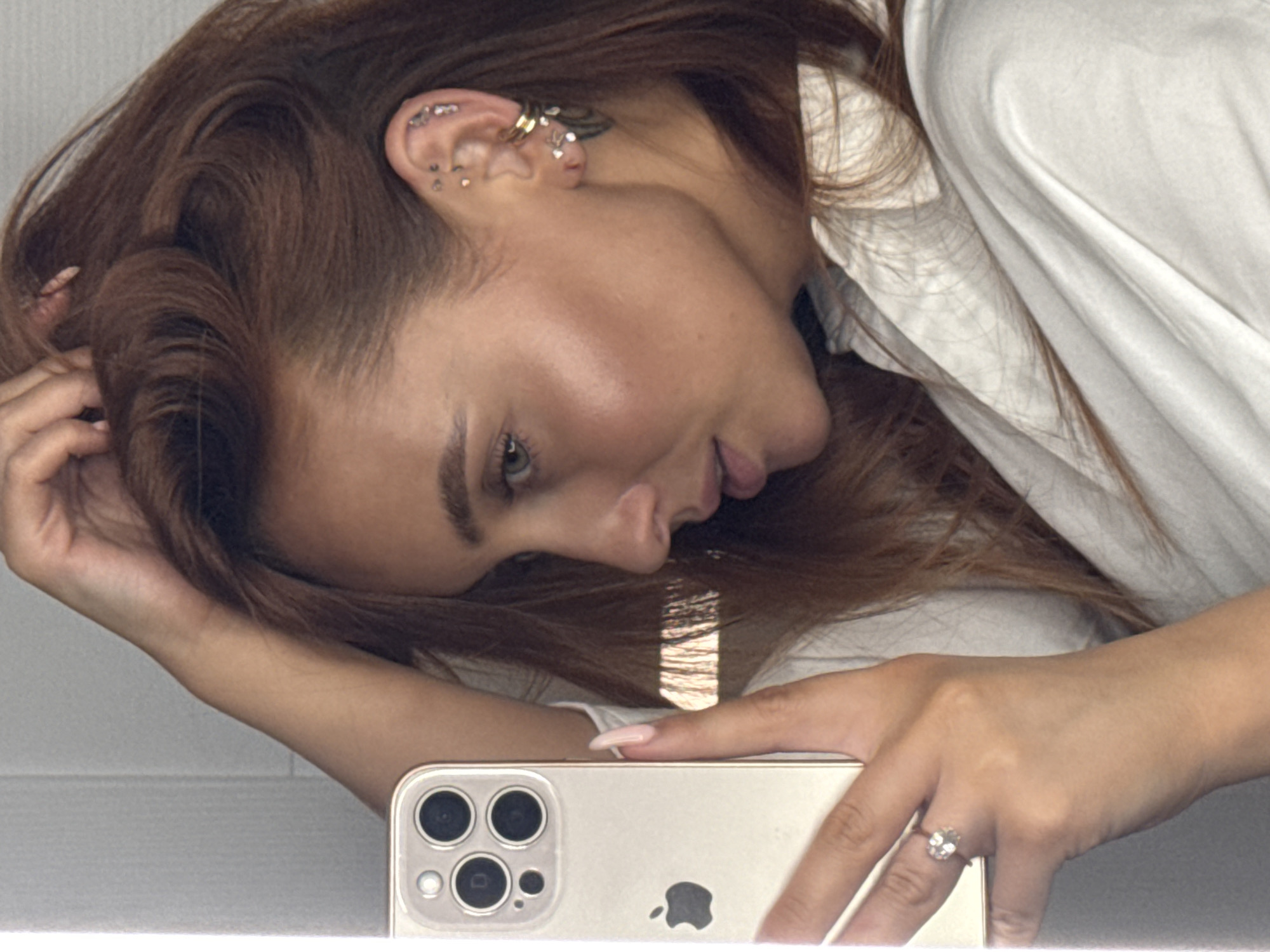 A close-up, side-profile shot shows a person holding a smartphone and their hand on their head, with their hair cascading over their shoulder. The person is wearing a white collared shirt and multiple earrings in their left ear. A ring with a large stone is visible on their left hand. The background appears to be a plain wall. There is no discernible information in the image that would allow for identification of the location in Chaikovsky, Russia.