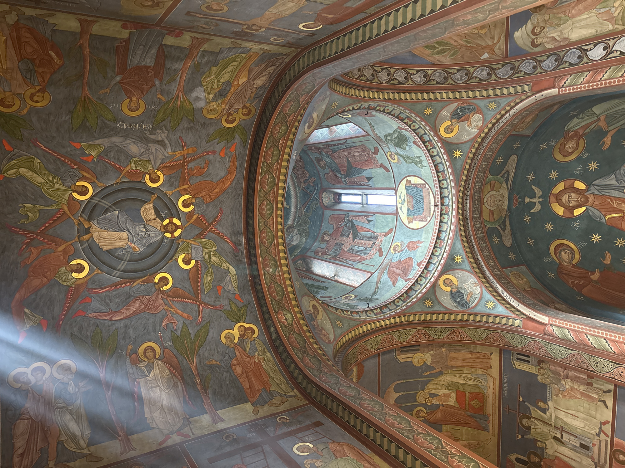 The image shows the interior of the Tbilisi Sioni Cathedral in Georgia. The view is an upward shot of the ceiling, which is covered with religious frescoes depicting various biblical figures and scenes. The artwork is intricate and colorful, with heavy use of gold halos around the figures' heads. A prominent beam of sunlight shines in from the left, illuminating a section of the frescoes.