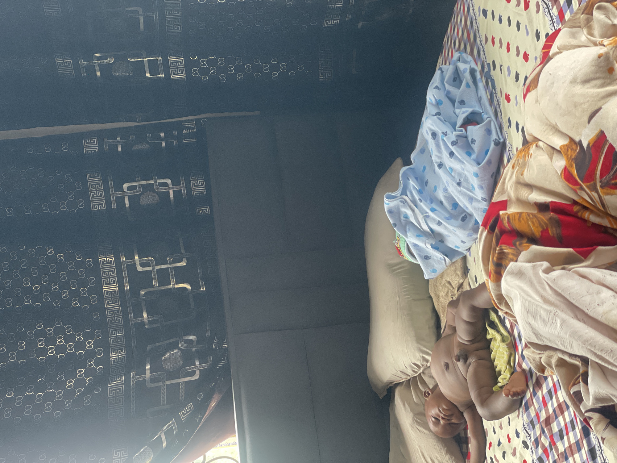 In Keffi, Nigeria, a baby sleeps on a bed in a room. The baby is lying on its back, with its legs spread and arms outstretched. The baby is naked except for a yellow cloth underneath its lower body. A light blue blanket with a pattern of whales is draped over the baby's legs. The bed is covered with a checkered comforter. In the background, a dark headboard is visible. The wall behind the bed is covered in wallpaper with a repeating pattern of interlocking geometric shapes and the letter "G" or "C" in a metallic sheen. The room appears to be lit by natural light coming from a window on the left side of the image.