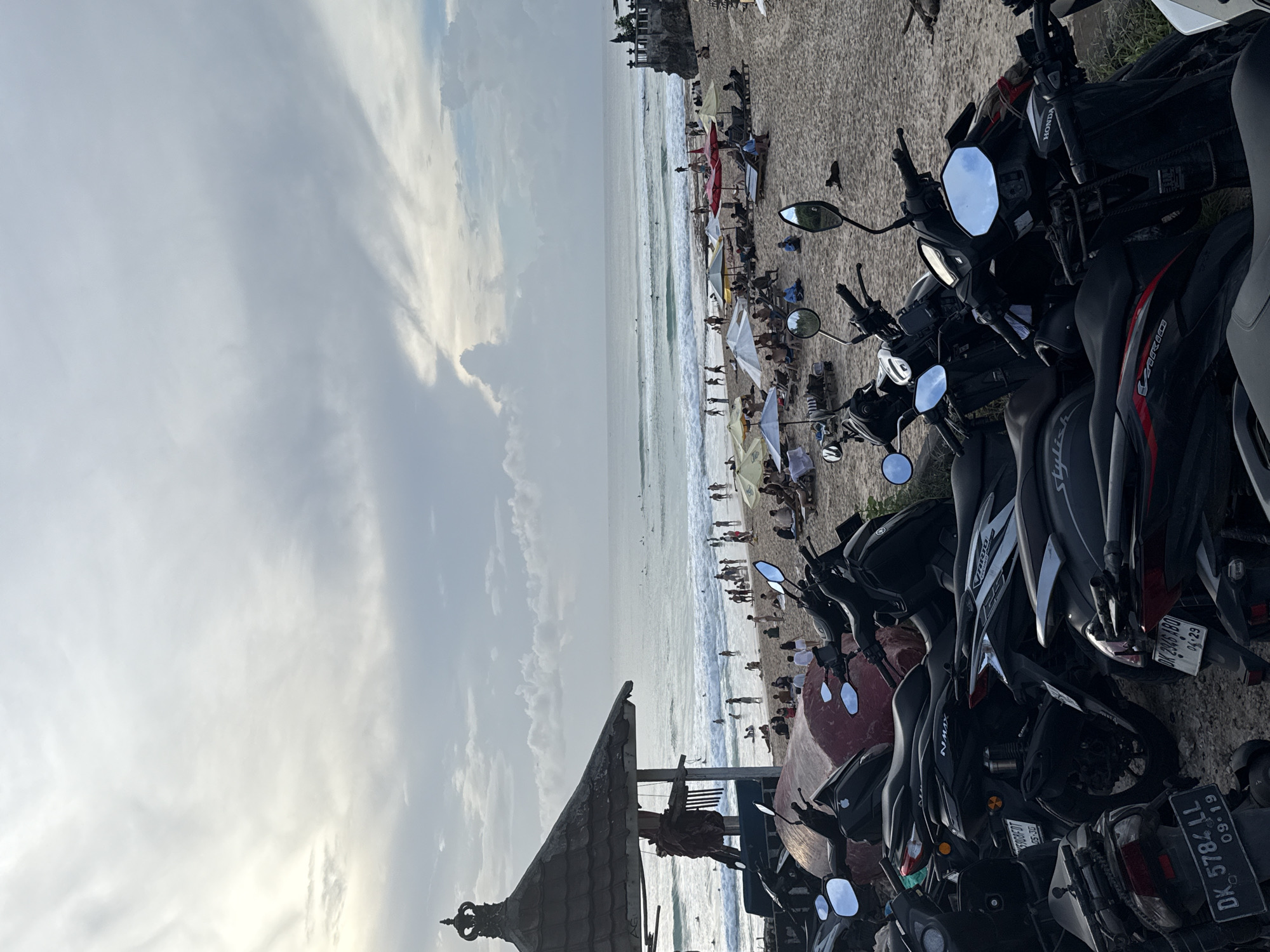 The image captures a vibrant beach scene in Kuta Selatan, Indonesia, teeming with activity under an overcast sky. In the immediate foreground, a line of black and dark-colored motorbikes, primarily scooters, are parked facing the beach, their rearview mirrors reflecting portions of the sky and surroundings. Several brand names such as "HONDA," "VARIO," and "Stylish" are visible on their bodies. License plates "DK 2045 ARO" (expiring 04-23), "DK 5784 LL" (expiring 09-19), and "DK 6700 FOT" (expiring 06-30) are readable on the rear of some bikes.
Beyond the parking area, a wide sandy beach stretches out, populated by numerous people. Many individuals are relaxing on beach chairs or directly on the sand, often shaded by a variety of beach umbrellas in red, white, yellow, and blue hues. Further towards the horizon, the ocean exhibits moderate waves, with several people visible in the water, likely swimming or surfing.
To the left, a traditional Balinese-style structure with an ornate, multi-tiered roof stands partially visible, suggesting a beachside vendor or facility. In the far distance on the left side of the beach, a rocky cliff or large natural formation rises from the sand, with some smaller structures atop it. The sky above is largely cloudy, with patches of lighter and darker gray, indicating a daytime setting, possibly late afternoon given the soft, diffused light. The overall atmosphere is one of leisure and recreation at a popular beach destination.