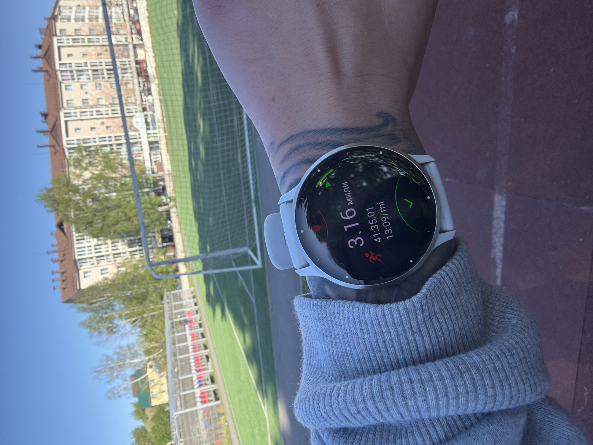 The image shows a person's wrist wearing a white smartwatch. The watch display indicates a running activity, with a distance of 3.16 miles and a time of 41 minutes and 35 seconds. The person is wearing a gray sweatshirt or long-sleeved shirt.
The background shows a sports field with green artificial turf and a soccer goal. In the distance, there is a large building with multiple floors. The setting appears to be outdoors on a sunny day.
Based on the architectural style of the building and available information, the location can be identified as being in Gorno-Altaysk, Russia. The large building in the background resembles apartment buildings or academic buildings found in the area.