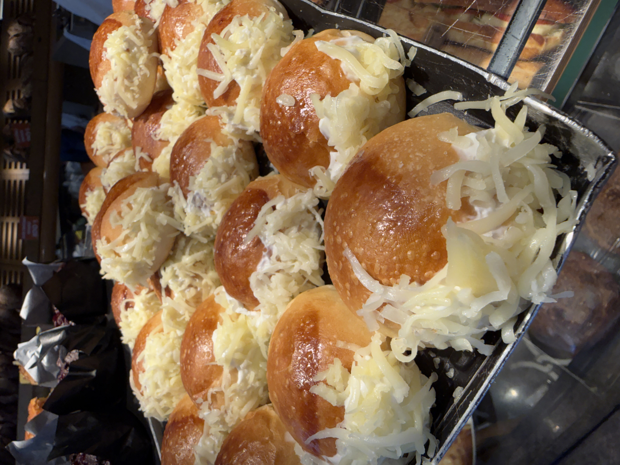 In Kadıköy, Turkey, a close-up shot captures a tray filled with freshly baked bread rolls generously topped with shredded white cheese. The rolls have a golden-brown, slightly glossy crust and are arranged closely together on a dark tray. The cheese appears to be finely grated and is scattered across the tops of the rolls, some of which have a white creamy filling peeking out from under the cheese. In the blurred background, there are other baked goods and what appear to be displays within a bakery or market setting.