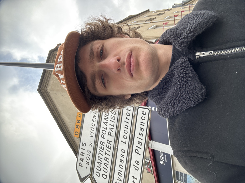 The image depicts a male individual in a close-up, chest-up outdoor portrait, set against an urban background in Joinville-le-Pont, France.
The subject is a young adult male with medium-length, curly brown hair. He wears an orange-brown baseball cap with white text, partially visible as "Ulv Res," worn forward. His facial features include a straight nose with a small dark spot on the right side, and full lips, with a neutral expression as he looks directly into the camera. He is dressed in a black jacket or coat featuring a dark grey, textured, sherpa-style collar and a visible silver zipper running down the front. A dark, possibly black, crew-neck garment is visible underneath the jacket.
Behind the individual, a white street signpost points to the left, displaying multiple destinations: "D 86 b" (a road designation), "PARIS - BOIS DE VINCENNES," "QUARTIER POLANGIS," "QUARTIER PALISSY," "Gymnase Lecuirot," and "Port de Plaisance." These signs identify the location as Joinville-le-Pont, a commune in the southeastern suburbs of Paris, France. Below the signs, a blue and white storefront sign for "EIFFAGE IMMOBILIER" is partially visible.
In the background, to the left, a light-colored building with architectural details like a cornice is visible. To the right of the subject, another multi-story, light-colored building with numerous windows and some shutters is present. A tall, slender, dark-colored pole extends vertically from behind the subject's head towards the top of the frame. The sky is overcast with light grey clouds.