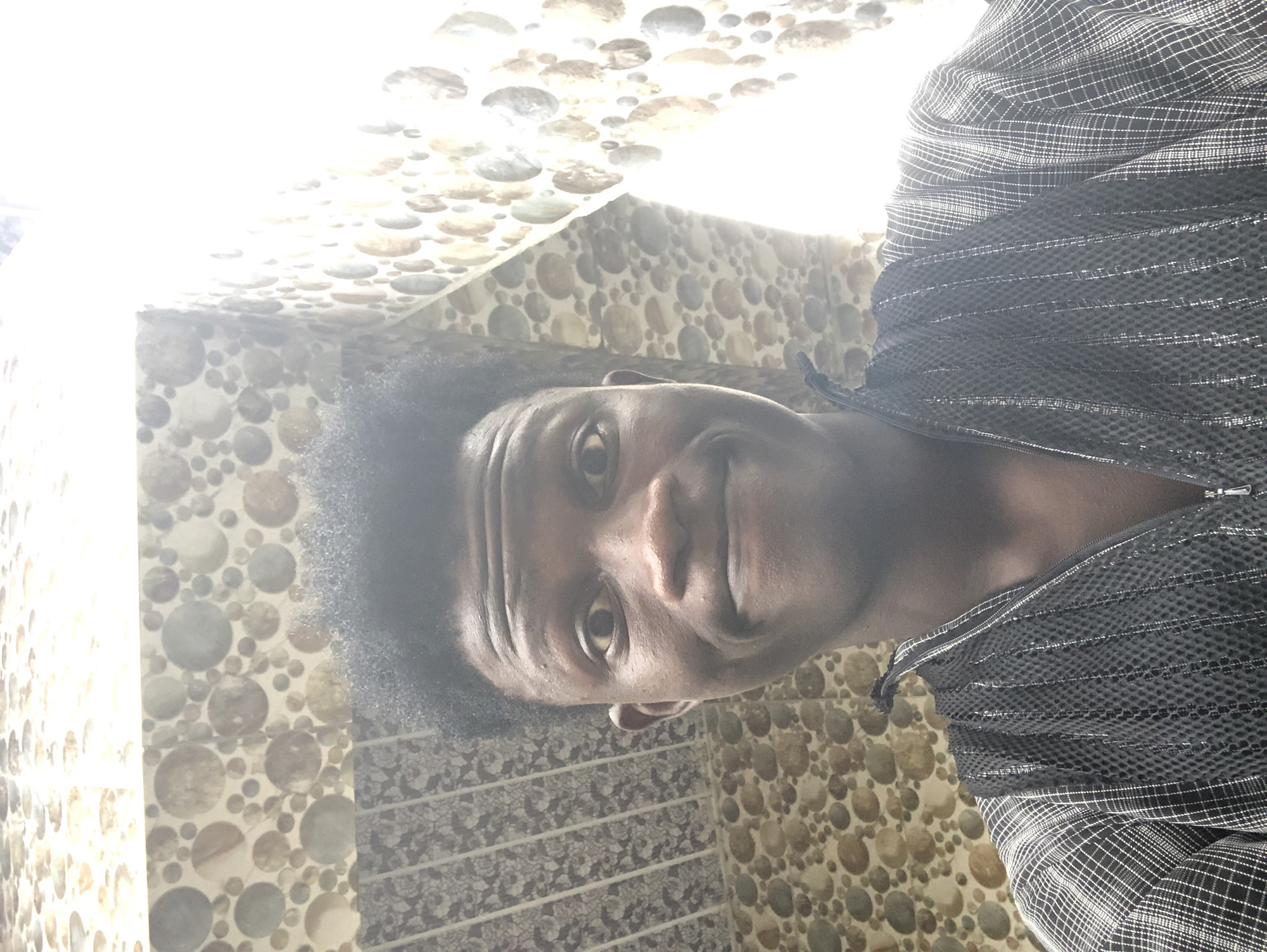 In Ado, Nigeria, a young Black male with dark, curly hair and dark skin is taking a selfie. He is wearing a black and white checkered shirt with a black vest layered over it. The vest has a zipper and a textured, patterned front. He is looking directly at the camera with wide eyes and a slight smile. The background appears to be a wall covered in a mosaic of circular tiles in shades of brown, beige, and grey. A bright light source is visible on the right side of the image, creating a glare. The composition is a close-up shot, focusing on the individual's face and upper torso.