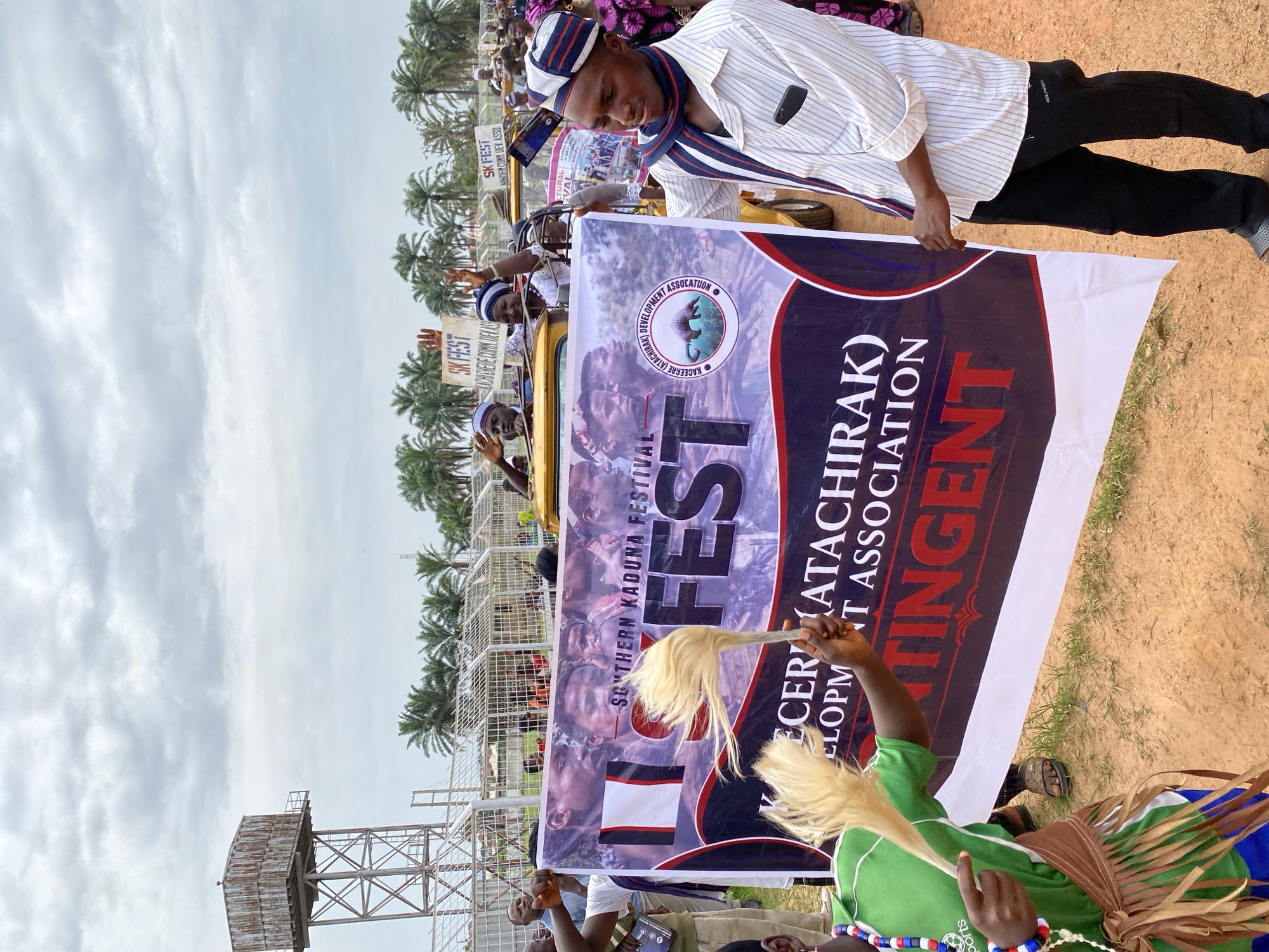 Captured in Kafanchan A, Nigeria, the image portrays a festive outdoor gathering. The focal point is a large banner proclaiming "SOUTHERN KADUNA FESTIVAL," also containing the words "KACEERRE (ATACHIRAK) DEVELOPMENT ASSOCIATION," and "FEST" in large font. People are holding the banner. One man, partially visible, wears a white and striped shirt with a scarf and a matching cap. A person in green stands in front of the banner, holding a white object resembling a whisk. Background features include palm trees and what appears to be a raised platform or structure. The sky is overcast.