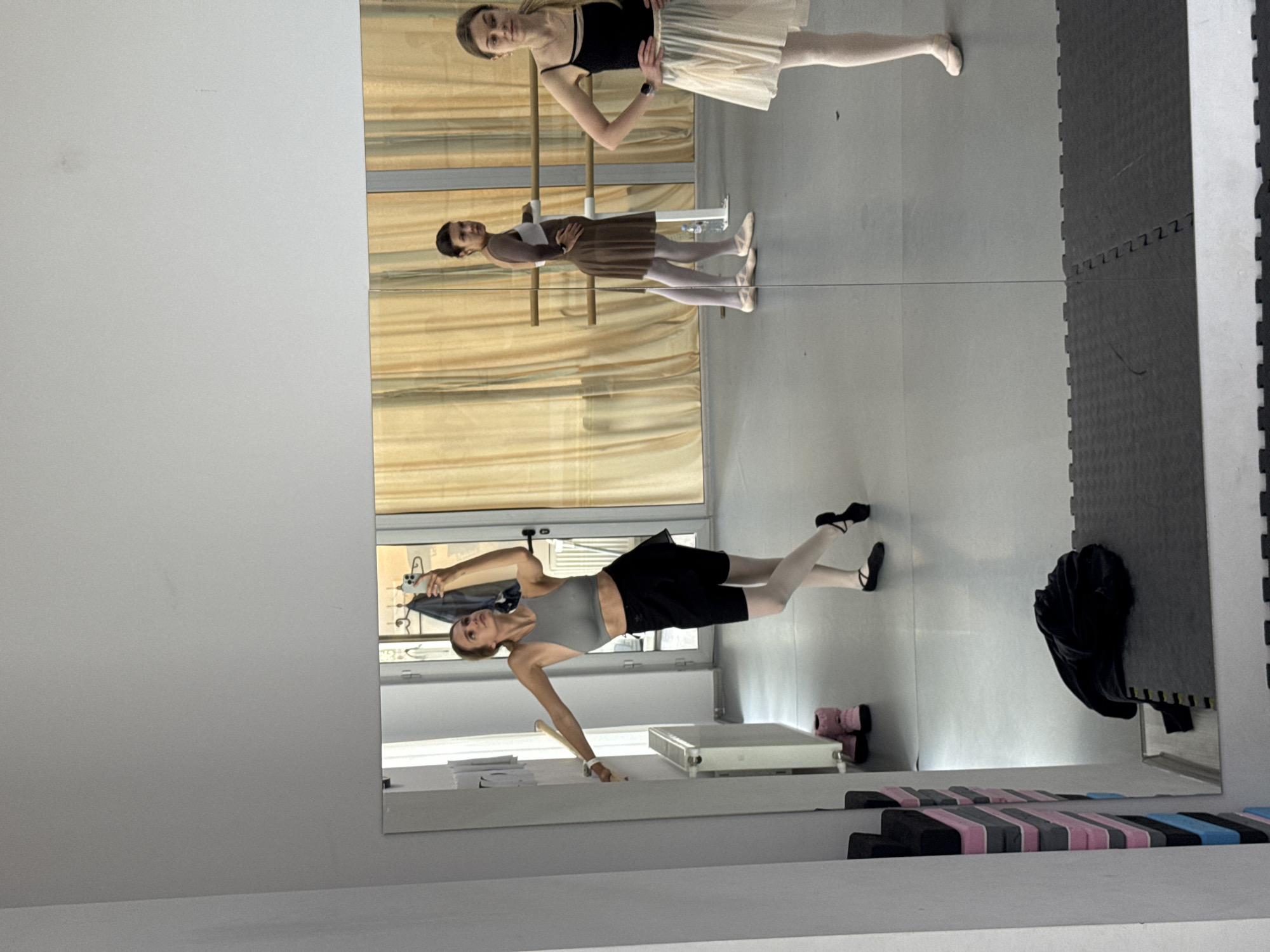 A ballet practice room is shown in the image. Three women are visible, either performing ballet or reflected in the mirror in the room. All three women are wearing ballet attire. One of the women is taking a picture of herself in the mirror. The backdrop features ballet bars, a large mirror, light-colored curtains, and workout mats.