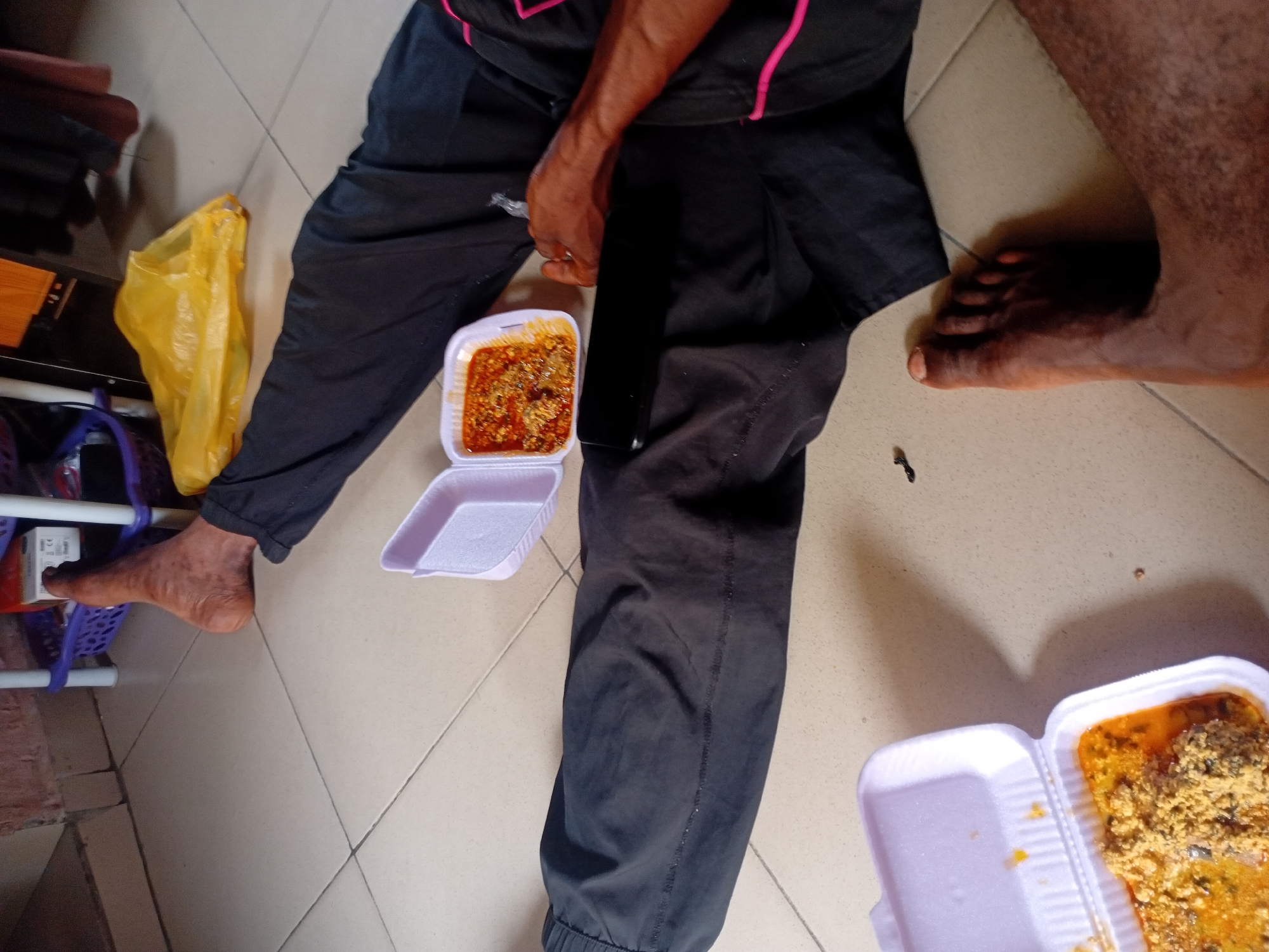 The image provides an overhead view of two individuals on a tiled floor, engaging in a casual meal, likely in Lokoja, Nigeria. One person is seen wearing black pants and a black shirt with pink trim, while the other's bare leg and foot are visible on the right.
Two white styrofoam take-out containers with open lids are placed on the floor between them. The container closer to the person in black pants holds a reddish, rich-looking stew or soup with chunks of what appears to be meat or fish. The second container, in the lower right, contains a yellowish-orange soup topped with a substantial amount of a light brown, grainy substance, consistent with Egusi soup, a common Nigerian dish made from ground melon seeds. The person in black pants appears to be holding a thin, metallic utensil, possibly a spoon or fork, near their food container.
Other objects visible include a yellow plastic bag on the left, a black smartphone lying on the floor next to one of the food containers, and parts of a purple plastic shelving unit in the background. The tiled floor and casual setup suggest an indoor, relaxed setting. There is no visible text in the image. The lighting indicates it is likely daytime.