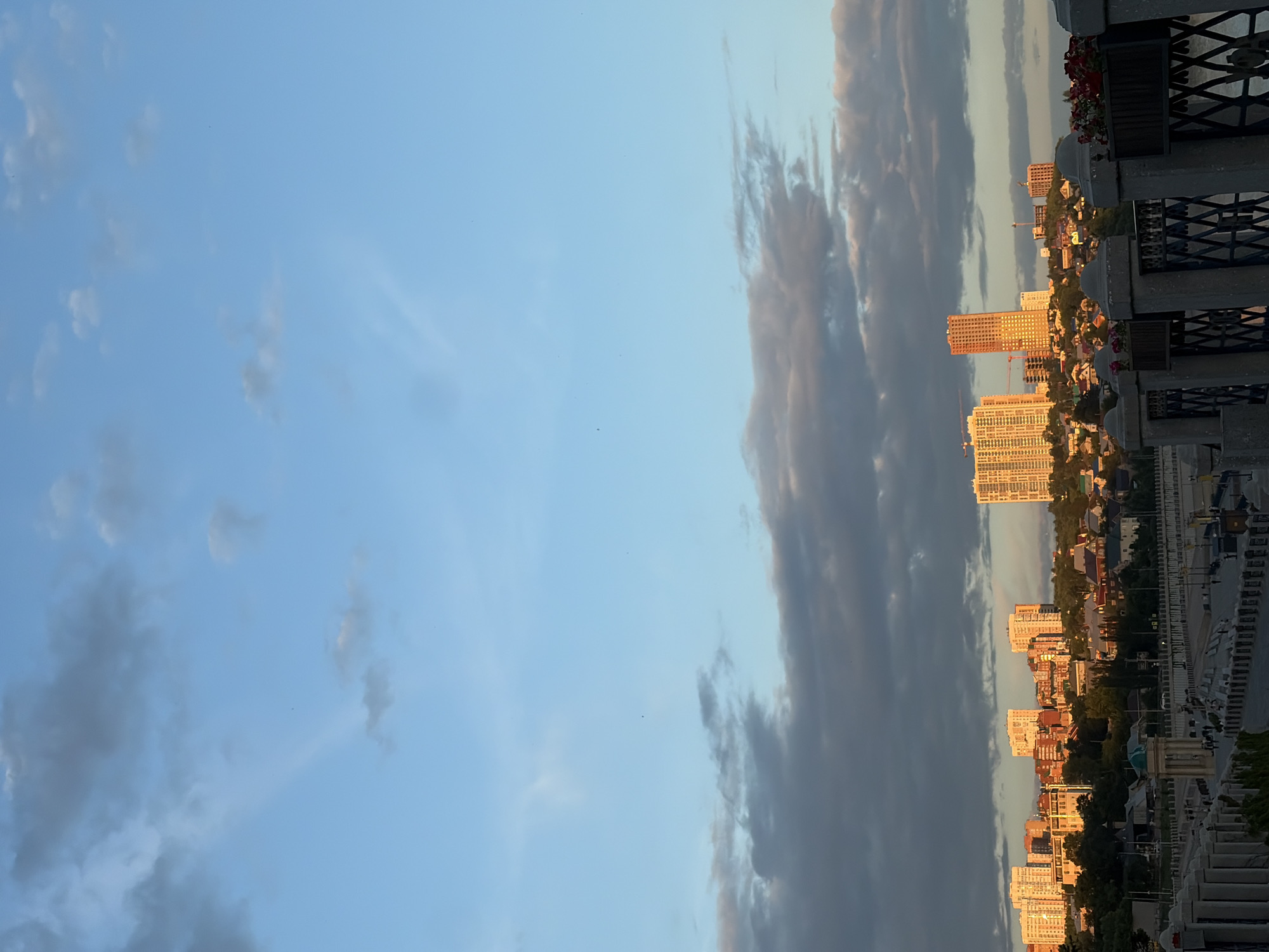 The image captures a cityscape of Ufa, Russia, likely at sunset, with a panoramic view of the embankment and the residential areas beyond. In the foreground, a stone embankment with decorative pillars and fencing runs along a body of water, possibly the Belaya River. Flower boxes with red flowers are visible on the fence. The middle ground shows a mix of houses and trees, with some architectural structures like a small domed building. In the background, several high-rise apartment buildings and some construction cranes dot the skyline, catching the warm light of the setting sun. The sky is a mix of blue and gray clouds, with some sunbeams breaking through.