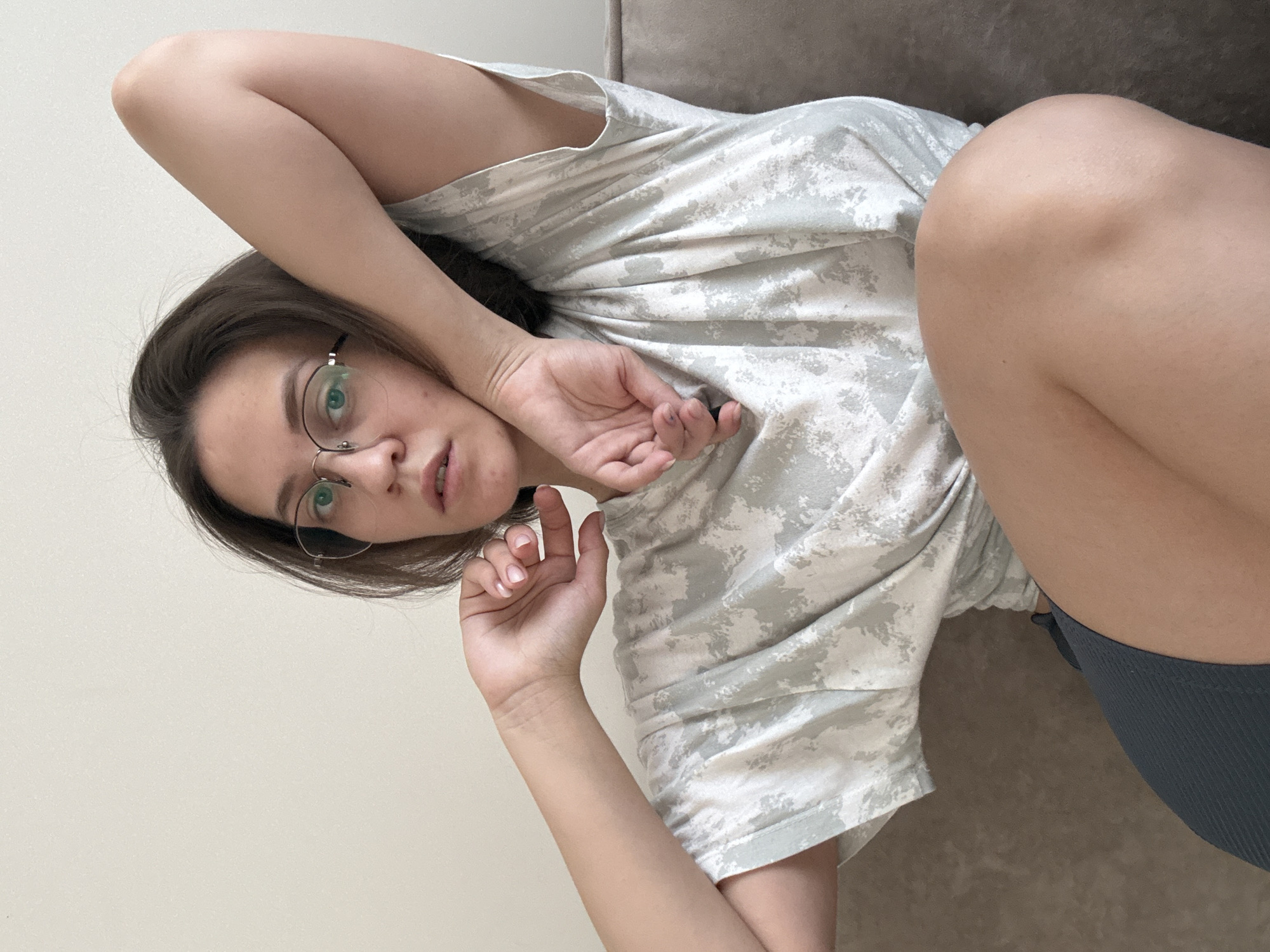 A woman with brown hair and round glasses is lying down, possibly on a sofa, in an indoor setting. She is wearing a light-colored patterned t-shirt and grey shorts. She has her arms raised. The background consists of a plain wall and the edge of what appears to be a sofa. There is no explicit information to connect the image to the city of Pervomaysky, Russia.