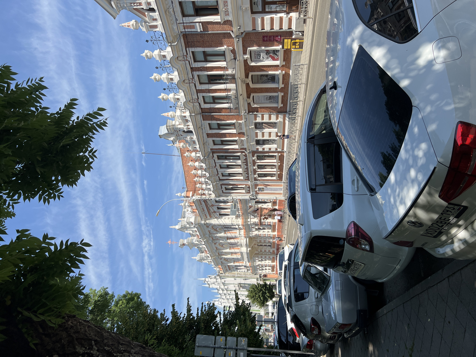 The image captures a street scene in Krasnodar, Russia, on a sunny day with scattered clouds. In the foreground, several cars are parked along the side of the road, casting shadows on the sidewalk. The vehicles include a white sedan, a white hatchback, and a silver sedan. A large tree with green leaves partially obscures the upper left portion of the frame.
The background features a row of ornate, multi-story buildings with distinctive architecture. These buildings are characterized by red brick facades, white decorative elements around windows and cornices, and numerous sculpted embellishments on the rooftops. A street lamp is visible above the buildings. A few individuals are walking on the sidewalk in front of the buildings. Several posters are displayed in the windows of one of the buildings, indicating it might be a museum or exhibition space. The street appears to be a typical urban thoroughfare with traffic lanes and a sidewalk.