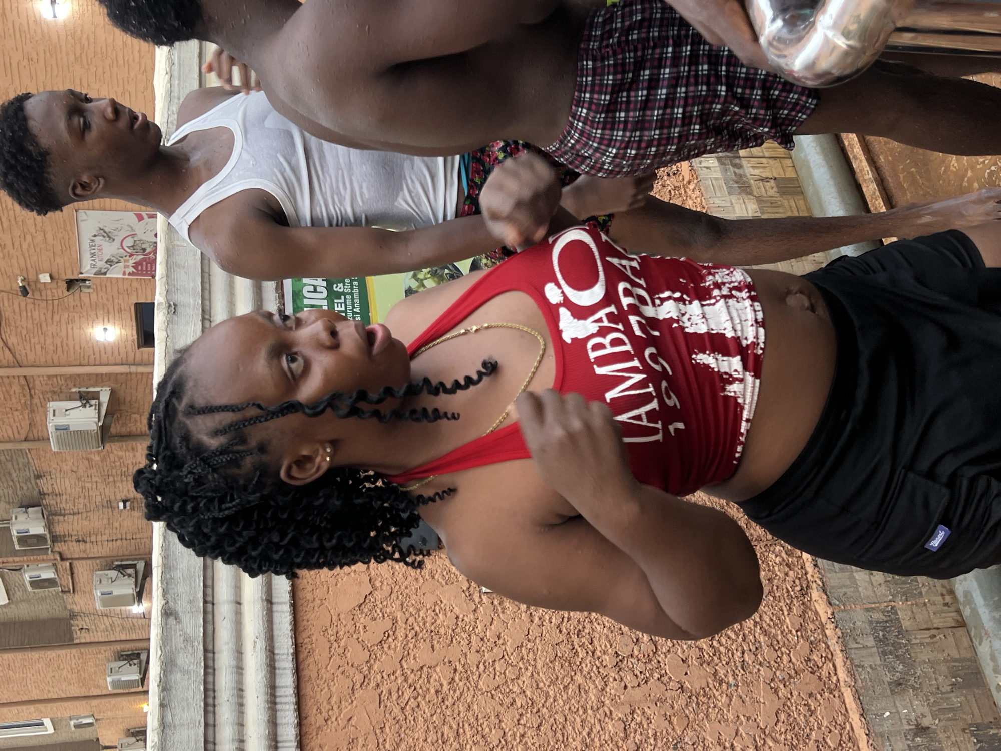 The image captures two individuals, a woman and a man, in an outdoor setting in Nkpor, Nigeria. The woman is in the foreground, looking animatedly towards the viewer with her mouth slightly open. She has dark, braided hair and is wearing a red sleeveless crop top with white text, black shorts, and a gold chain around her neck. Her arms are slightly raised, suggesting she is mid-action, possibly dancing or reacting to something.
Behind her, partially visible, is a man wearing a white tank top and red and black plaid shorts. Both individuals appear wet, implying they have been in water or are sweating, consistent with a lively activity or warm climate.
The background features a light brown, textured building wall adorned with multiple white air conditioner units. A white poster on the wall displays the text "FRANKVIEW KITCHEN." Below it, another partially obscured sign shows text including "LICA," "EL &," "zurumu Stre," and "si Anambra," providing a strong location cue. A shiny, silver metal object is visible in the upper right. The ground beneath them is a mix of concrete and a rougher, possibly earthy texture.
Visible text on the woman's red top includes "LAMBAO" and "1970/1971". A small label on the man's shorts reads "Vinculo." The scene suggests a casual social gathering or event during the daytime.