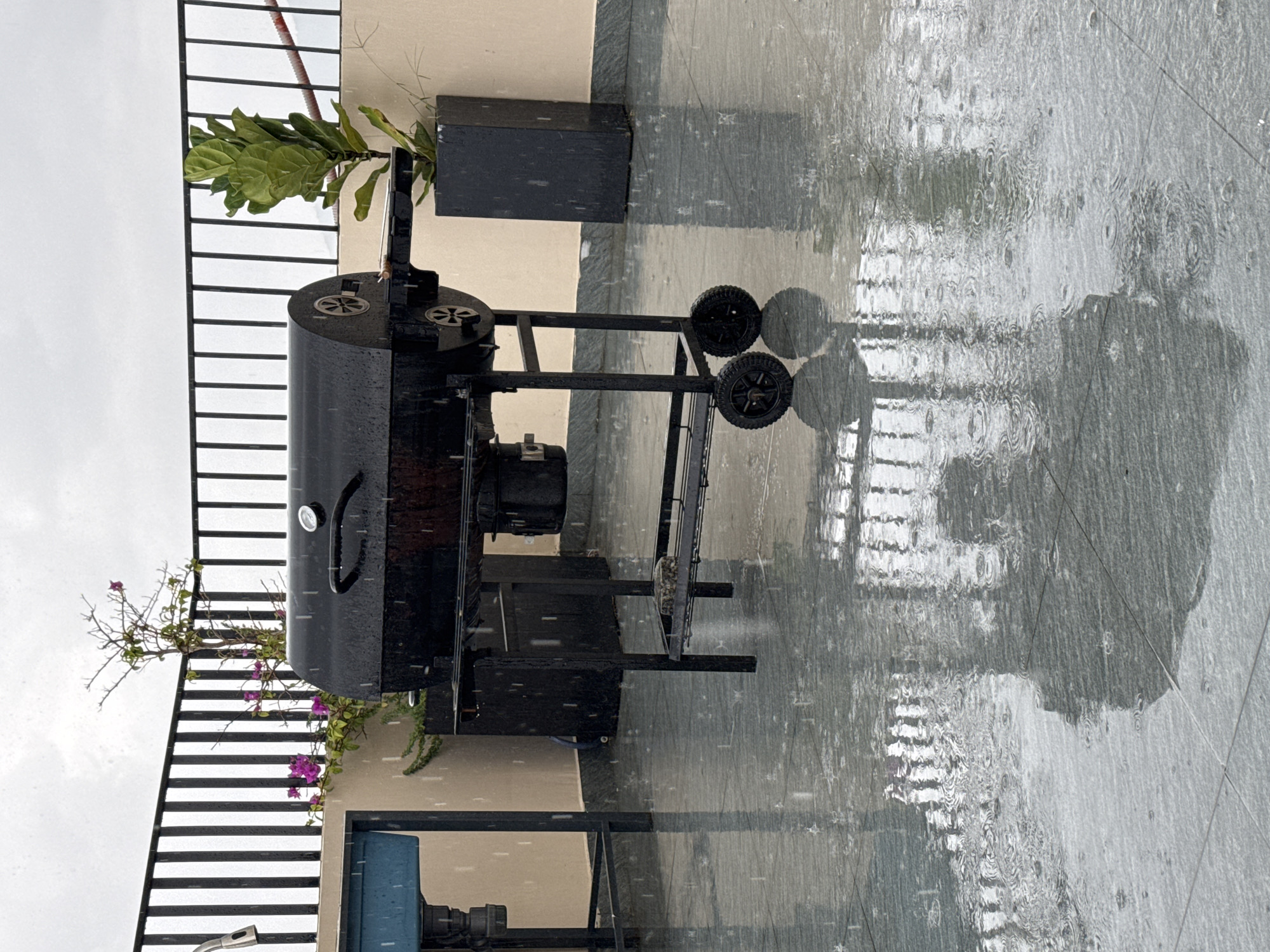 The image captures a black grill situated on a tiled patio during a rain shower. The grill is the central focus, with its wheels, shelves, and lid clearly visible. The patio floor is wet and reflective, showing ripples from the falling raindrops. In the background, a black metal railing encloses the patio, and a plant in a dark planter is visible to the right of the grill. To the left, a blue basin or sink is partially visible. The sky is overcast and grey, typical of rainy weather. The scene suggests an outdoor area, possibly a rooftop or balcony, in an urban setting, consistent with areas in Đà Nẵng, Vietnam, where such outdoor spaces are common. No people or specific identifiable landmarks of Đà Nẵng are present, only general elements of an urban outdoor environment.