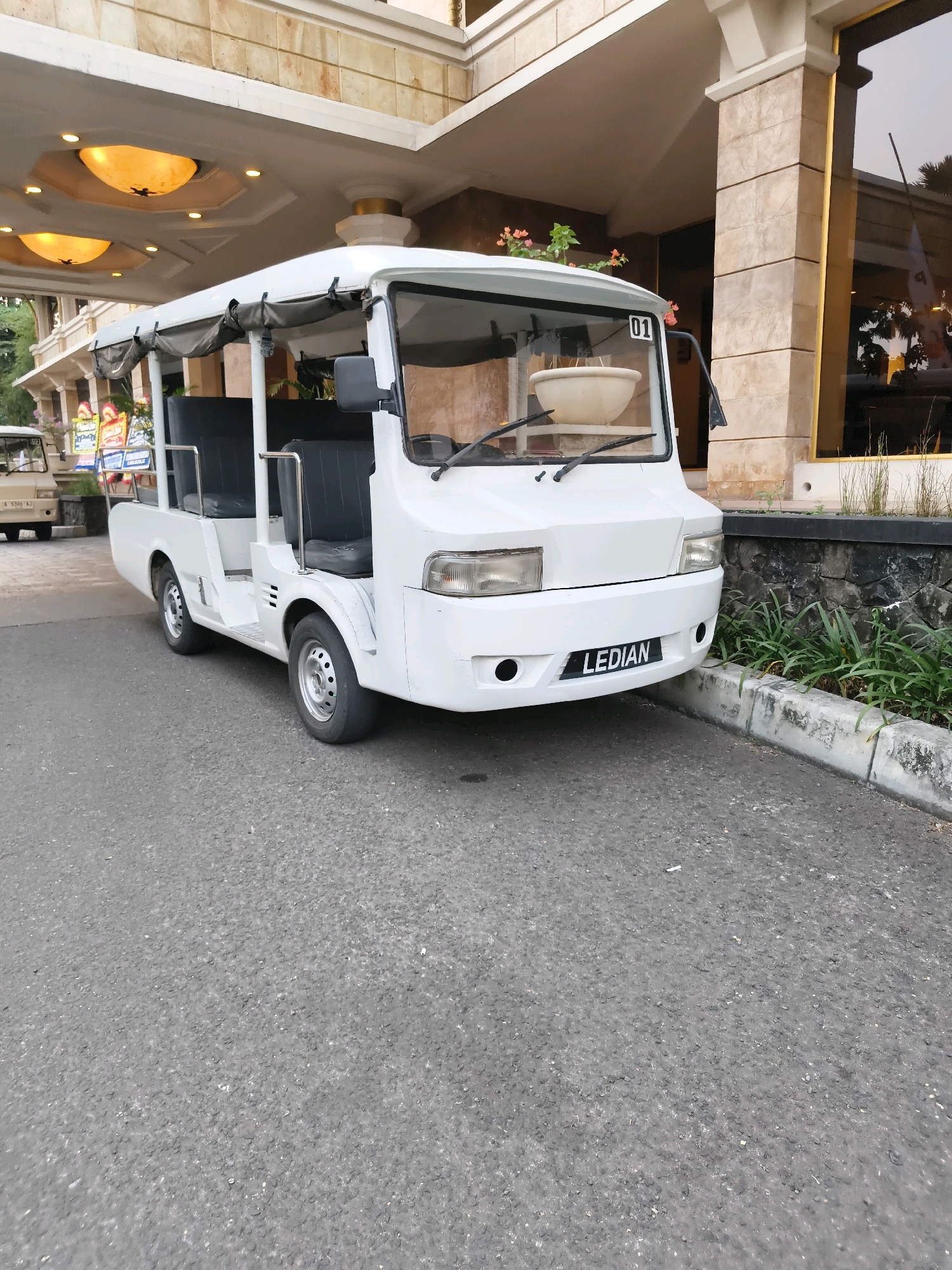 A white, open-air shuttle bus with a black roof is parked on an asphalt road in front of a building with light-colored stone cladding and a covered walkway with decorative lighting. The bus has two rows of black seats and a "LEDIAN" license plate on the front. A white flower pot sits on the dashboard. In the background, there are some green plants and a glimpse of another vehicle. The image appears to have been taken during the day.