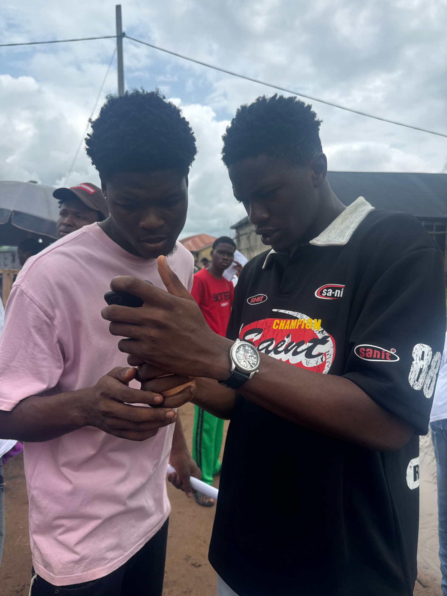 Two young men are standing close together in what appears to be an outdoor, possibly urban, setting. The man on the left wears a light pink t-shirt and is holding a dark-colored mobile phone, looking down at it. His hands are intertwined with the hands of the man on the right. The man on the right wears a black collared shirt with white lettering and logos. On his left wrist, he wears a silver watch with a black leather strap. He is also looking down, seemingly at the phone or something in the hands of the other man.
In the background, other individuals are visible. One person in a red t-shirt can be seen further back. The ground appears to be dirt or unpaved road. Overhead, there are wires and a cloudy sky. The scene suggests a casual, everyday interaction in a community. This specific location is identified as Ihiagwa, Nigeria.