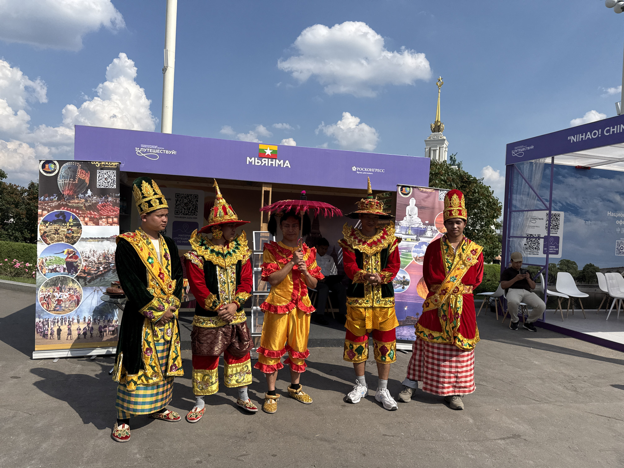The image depicts an outdoor exhibition in Moscow, Russia, likely at VDNKh (Exhibition of Achievements of National Economy), given the distinctive architecture visible in the background. A group of five individuals dressed in traditional Myanmar attire stand in front of a purple exhibition booth. The attire consists of elaborate headdresses, brightly colored tunics with gold embellishments, and patterned skirts or pants. The person in the center holds a decorative pink umbrella. Behind them, a banner displays the text "МЬЯНМА" (Myanmar) with the Myanmar flag. To the right, another banner reads "'NIHAO! CHIN..." indicating a China-related exhibit. In the background, a tall building with a star-tipped spire, characteristic of VDNKh, is visible under a bright blue sky with scattered white clouds. Several white chairs are arranged near the exhibition booths, with a person sitting and looking at a phone in the far right. The ground is paved asphalt.