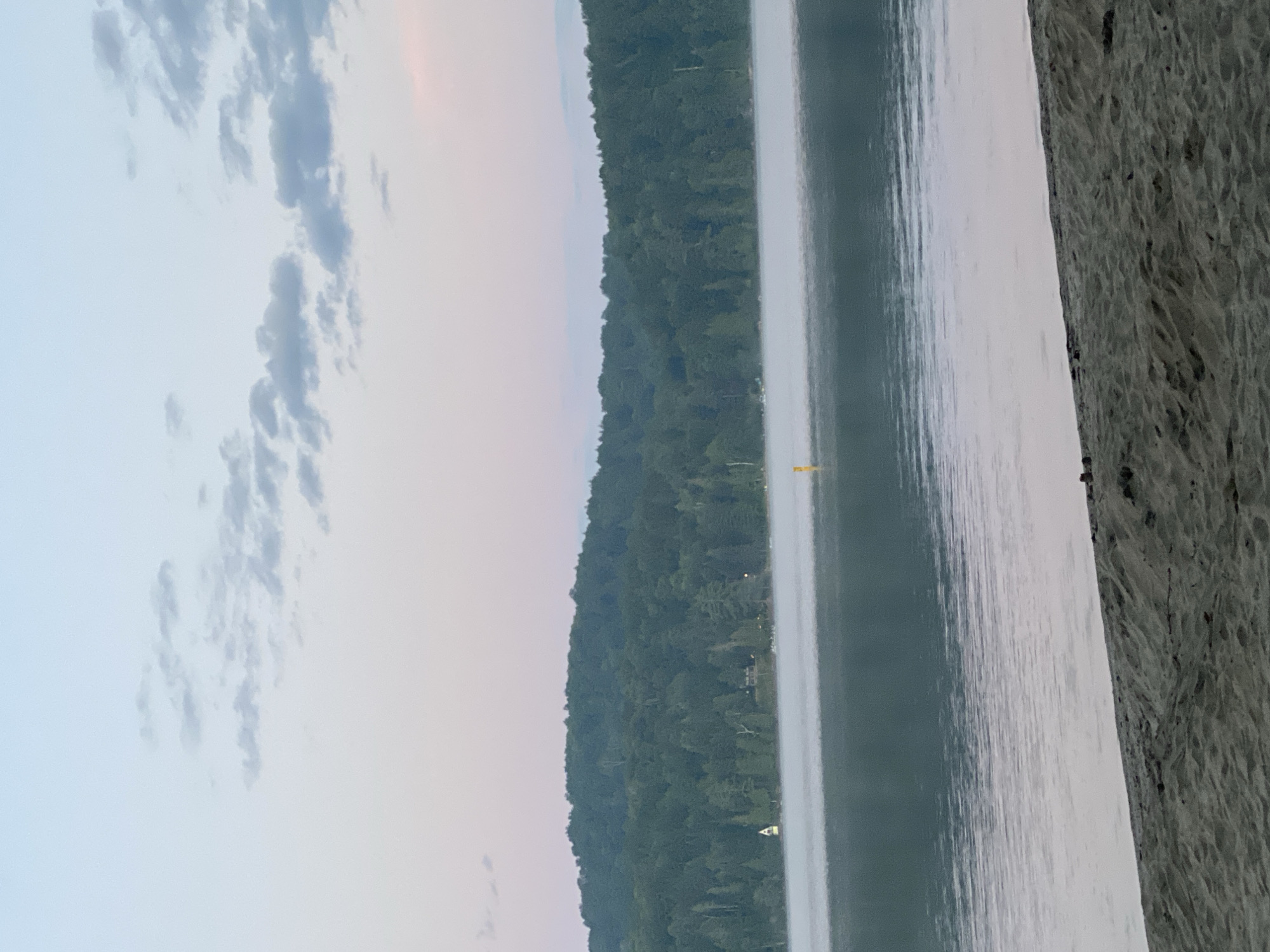 This is a photograph of a serene lake scene, likely at dawn or dusk, with a sandy beach in the foreground. The water is calm, with a gentle mist rising from its surface. Beyond the lake, a dense forest covers a rolling hillside. The sky is a soft gradient of pale blue and pink, with a few wispy clouds. A small yellow buoy is visible in the water, and a sailboat with a white sail can be seen near the shore on the left side of the image. The beach is covered in sand, with some scattered debris. The overall atmosphere is peaceful and natural. This location is likely in Duhamel, Canada, given the natural landscape and typical weather conditions for the region.