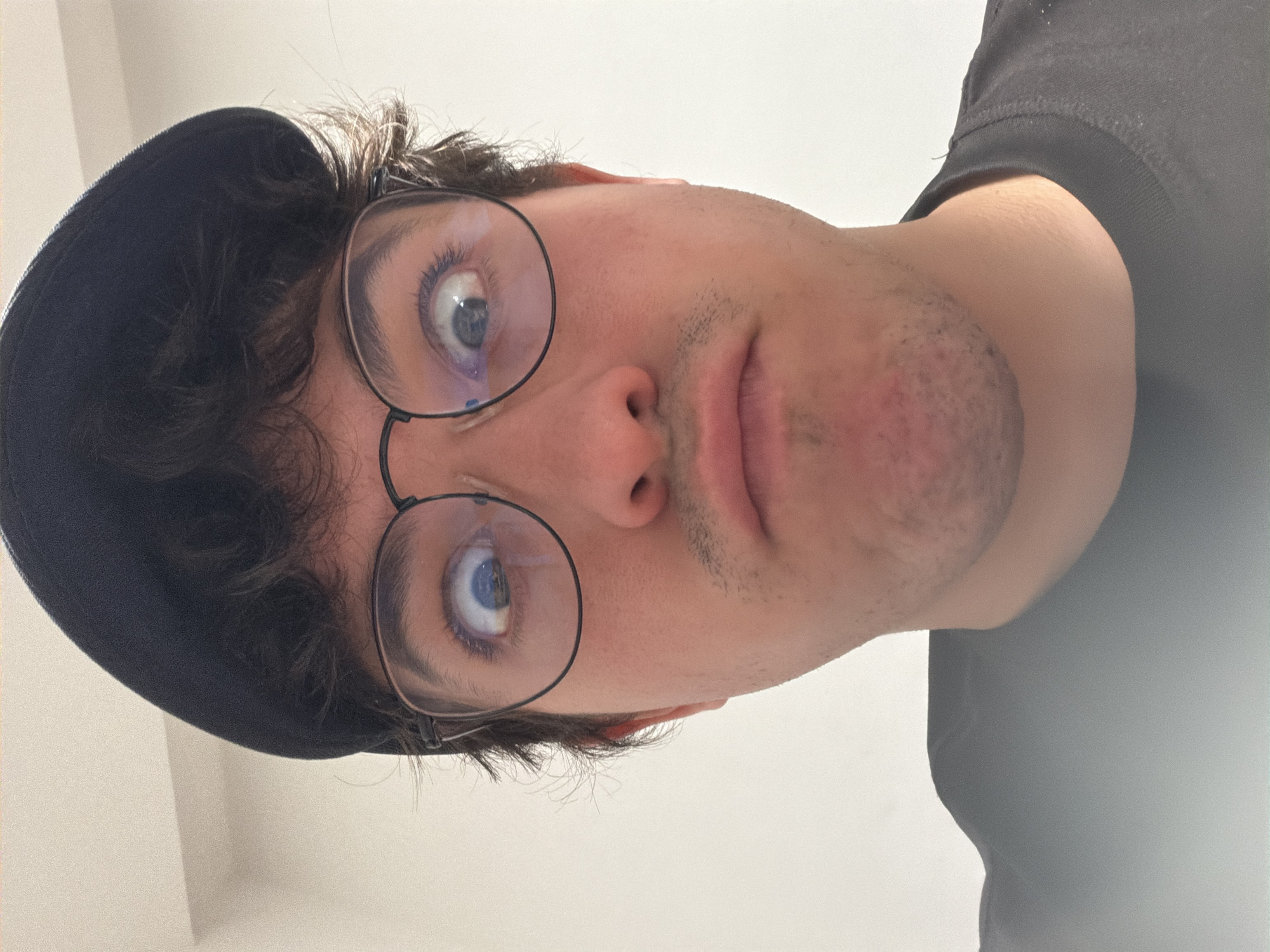 The image is a close-up, low-angle selfie of a young person wearing glasses and a dark cap. The person has brown, curly hair and is looking directly at the camera with wide, light-colored eyes. A light stubble is visible on their chin and upper lip. They are wearing a dark t-shirt. The background is a plain, light-colored wall, suggesting an indoor setting. The lighting is bright, indicating it could be daytime. The image is oriented vertically, as if taken from below the subject's head. No text is visible. The location context of Cancún, Mexico, is not directly discernible from the visual elements of the image itself.