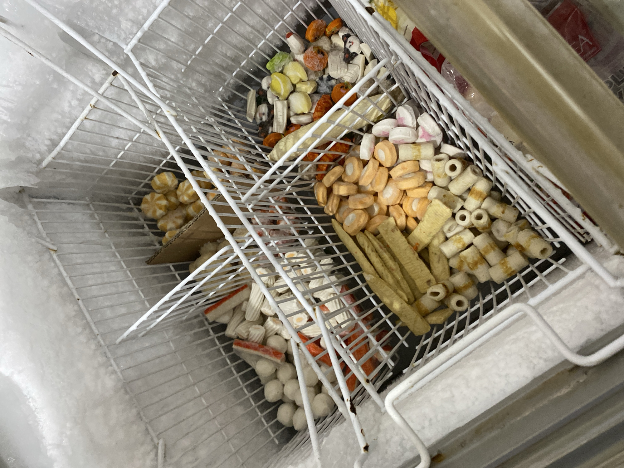 The image shows the interior of a freezer in Payatieng, Indonesia, filled with various frozen food items. The freezer is a wire-rack style, with multiple shelves containing a mixture of seafood, vegetables, and other processed frozen foods. Items include what appear to be fish cakes, imitation crab sticks, small white meatballs, and mixed frozen vegetables such as peas and corn. Some packages are visible in the background, including one with red packaging and text. The freezer walls are heavily frosted, indicating it is functioning but likely requires defrosting. The overall scene depicts a typical home or commercial freezer stocked with frozen ingredients.