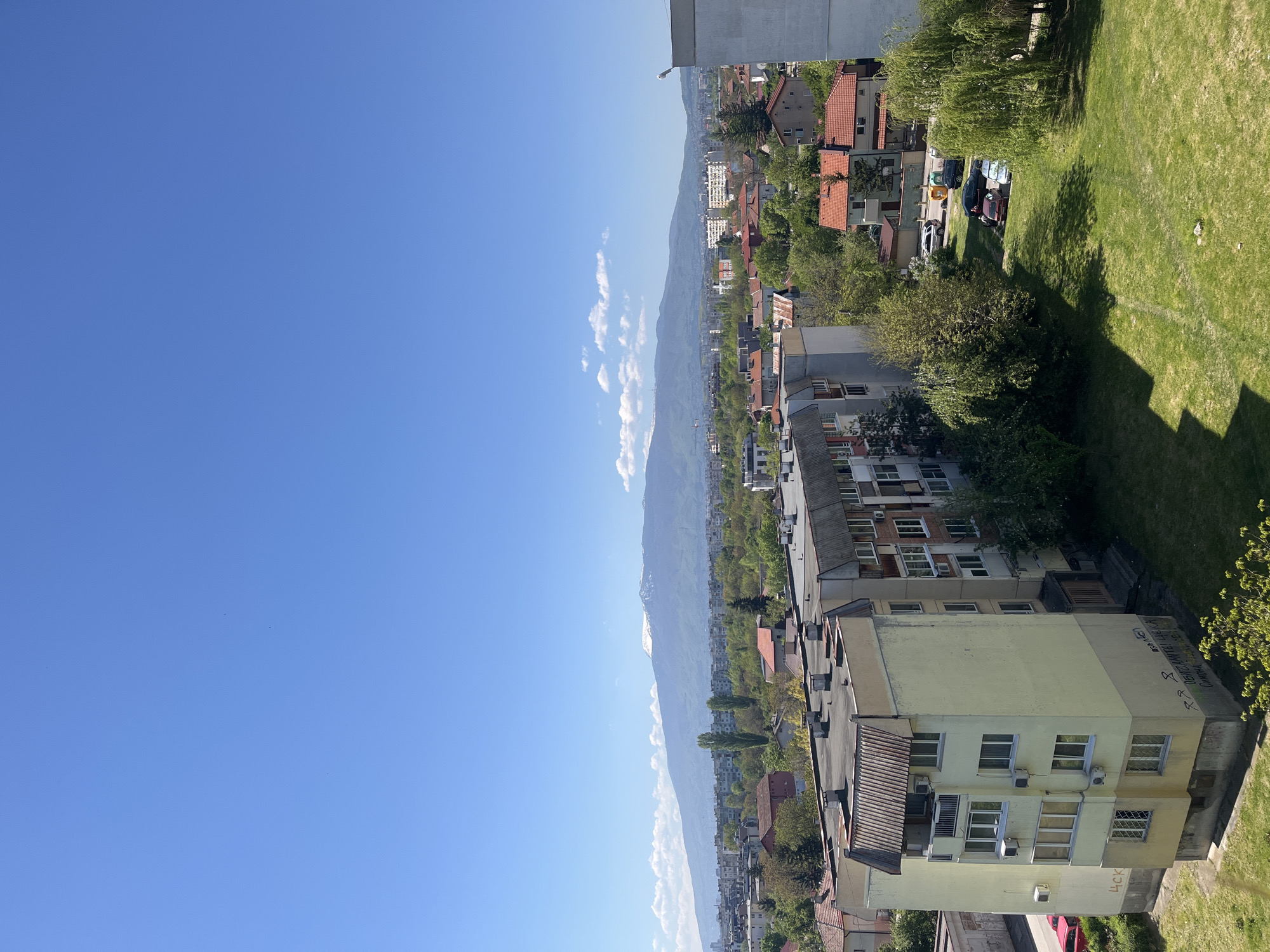 The image shows a view from a high vantage point, likely a building, overlooking a neighborhood in Sofia, Bulgaria. The sky is clear and blue, occupying a large portion of the left side of the image. A mountain range is visible in the background, partly obscured by a line of scattered clouds.
The cityscape comprises a mix of residential buildings, ranging from single-family houses with red tile roofs to taller apartment buildings. Green trees and lawns intersperse the built environment, suggesting a residential area. Several cars can be seen parked in the area, indicating a typical suburban setting.
The nearest building appears to be a multi-story apartment building. The color of the building is light green, and there is graffiti visible.