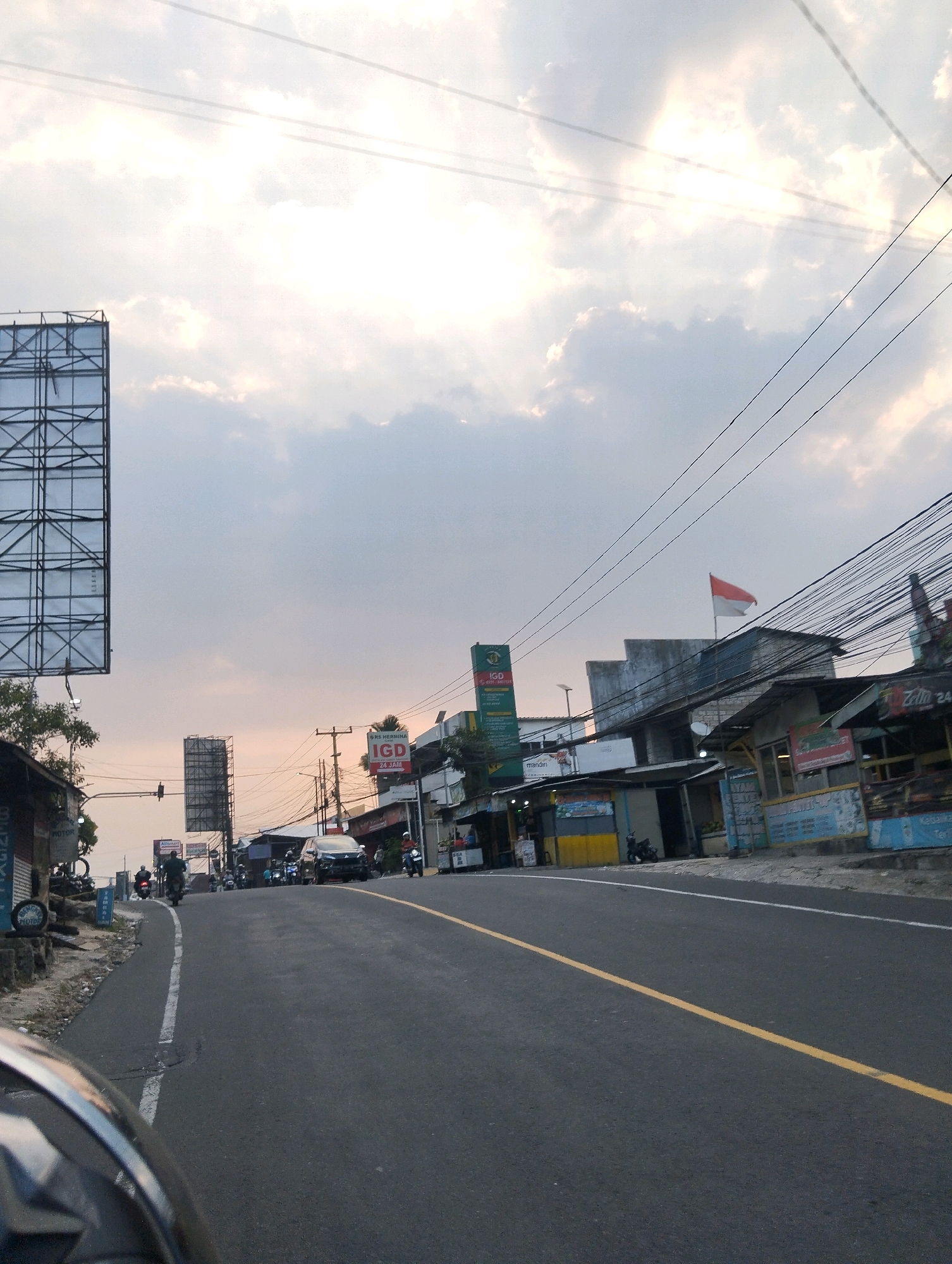 This is a photograph taken from a motorcycle at eye-level, looking down a two-lane road with a yellow center line. The road curves to the right and is lined with buildings on both sides, suggesting an urban or semi-urban environment. The buildings appear to be small shops or businesses, with some displaying signage. One sign, prominently visible, indicates "IGD 24 JAM," which translates to "Emergency Room 24 Hours," suggesting a medical facility nearby. Another sign, partially obscured, reads "RS HERMINA," likely referring to a hospital.
On the left side of the road, a large, empty billboard structure stands tall against a cloudy sky. A few trees are visible near the billboard. On the right side of the road, a red and white flag, likely the Indonesian flag, is flying from the roof of a building. There are several motorcycles and a few cars on the road, with a few people visible riding the motorcycles. The sky is overcast with patches of sunlight breaking through, indicating it might be late afternoon or early morning. The overall impression is a bustling street scene in Indonesia, possibly in the city of Megamendung, given the potential presence of "RS HERMINA" which is a hospital chain with branches in Indonesia. The weather appears to be mild, and the road surface is asphalt. There are numerous power lines crisscrossing the scene, typical of urban infrastructure in Indonesia.
