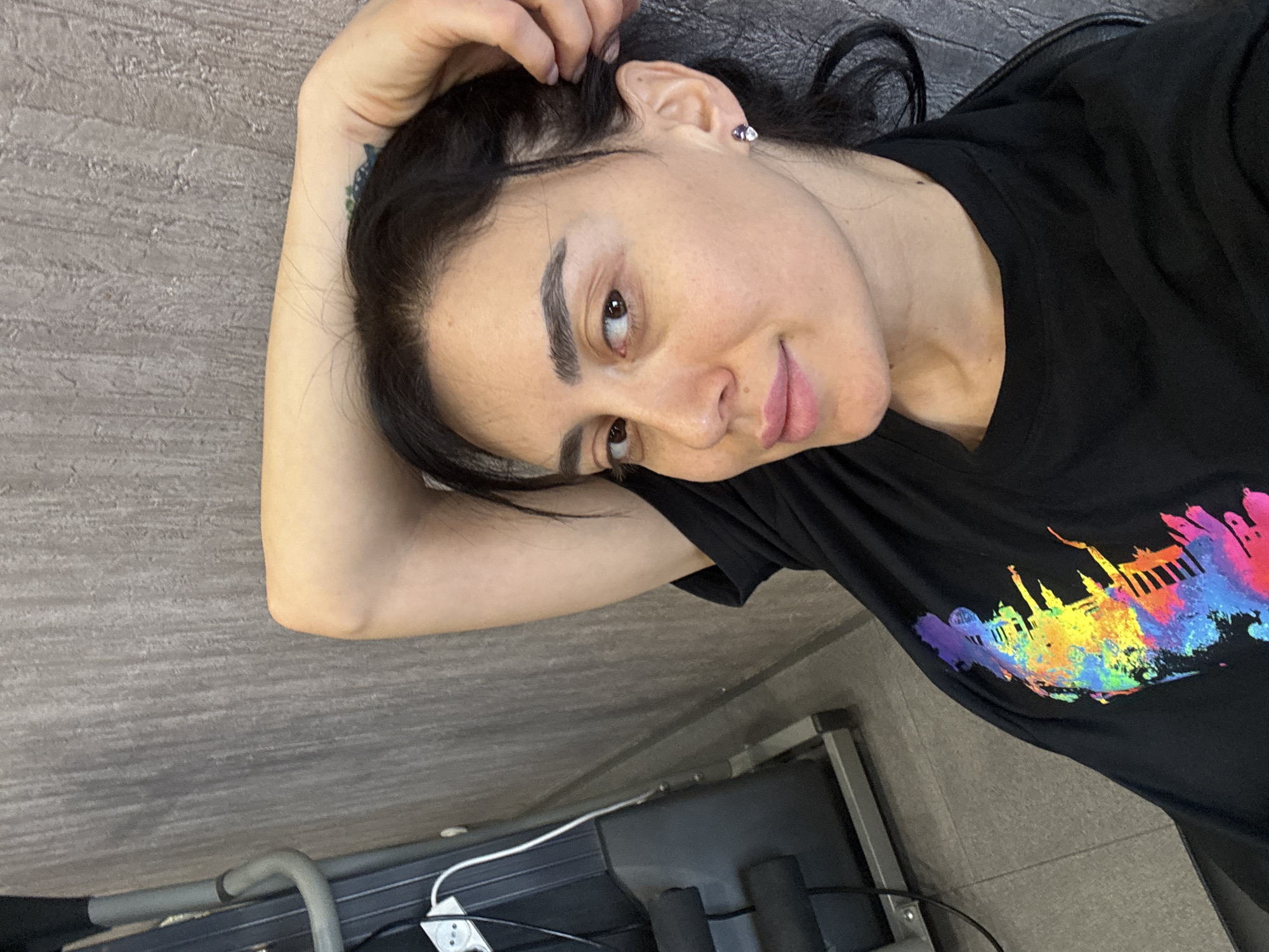 A woman lies on a tiled floor next to a gray exercise machine with a white power cord. She rests her head on her left hand, fingers touching her hair. A small tattoo is visible on her left wrist. She is wearing a black t-shirt with a colorful graphic of the Tbilisi skyline on it. The woman is looking towards the camera with a slight smile. A small stud earring is visible in her left ear.