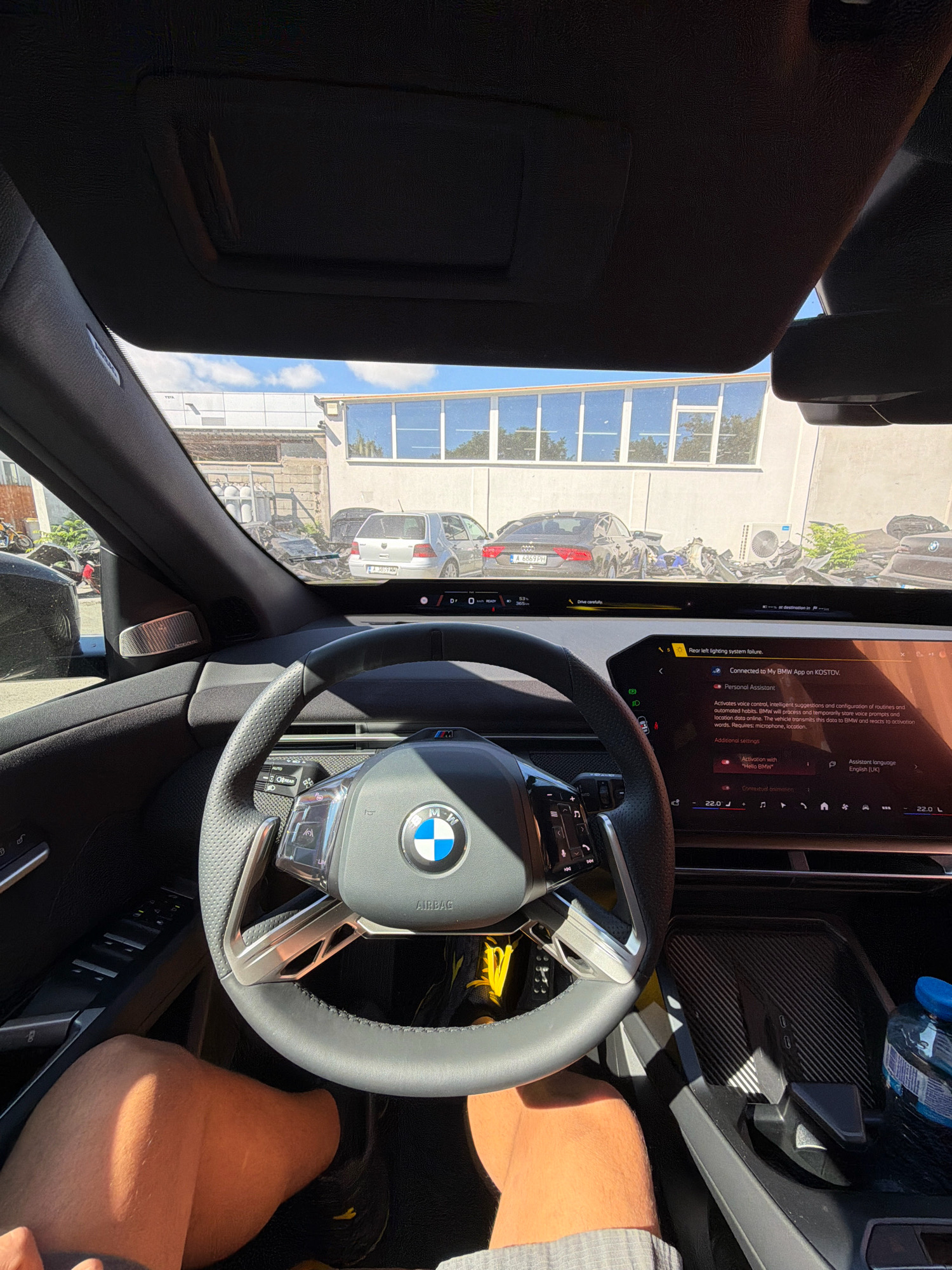 The image shows the interior of a car, looking out at a street scene in Burgas, Bulgaria. A person's legs are visible in the foreground, with the car's steering wheel and dashboard in the center. Through the windshield, several cars are parked on the street, including a silver Volkswagen Golf and a black Audi. A building with large windows is visible in the background. The sky is blue with some clouds. The overall impression is of a sunny day in a city.