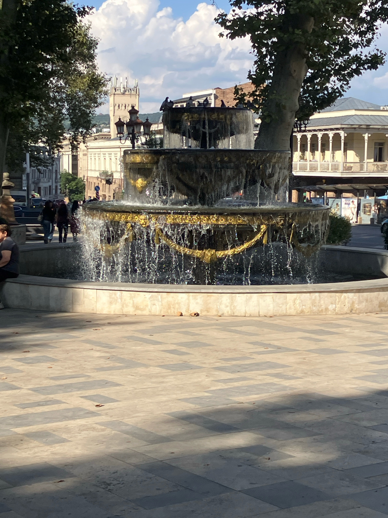 The image depicts a multi-tiered ornate fountain with gold accents in the foreground, with water cascading down its levels. The fountain is situated on a paved plaza with a checkered pattern of light and dark tiles. Behind the fountain, a large tree trunk is visible, its branches partially obscuring the sky and buildings.
In the background, several buildings are visible. To the left, a tall, tower-like structure with decorative architectural elements and modern-looking antennae on top stands prominently. Adjacent to it, a long, light-colored building with a colonnaded facade is seen. Further to the right, another building with a covered balcony and ornate railings is present.
Several people are walking in the mid-ground, near the fountain. One person is seated on a bench to the far left of the frame. Pigeons are perched on top of the fountain and on ledges of the background buildings. The scene appears to be outdoors, in a public urban space, under a bright sky with scattered clouds. The overall impression is that of a sunny day in a European city.
This location is the Fountain of Peace on Rustaveli Avenue in Tbilisi, Georgia, in front of the Parliament of Georgia.