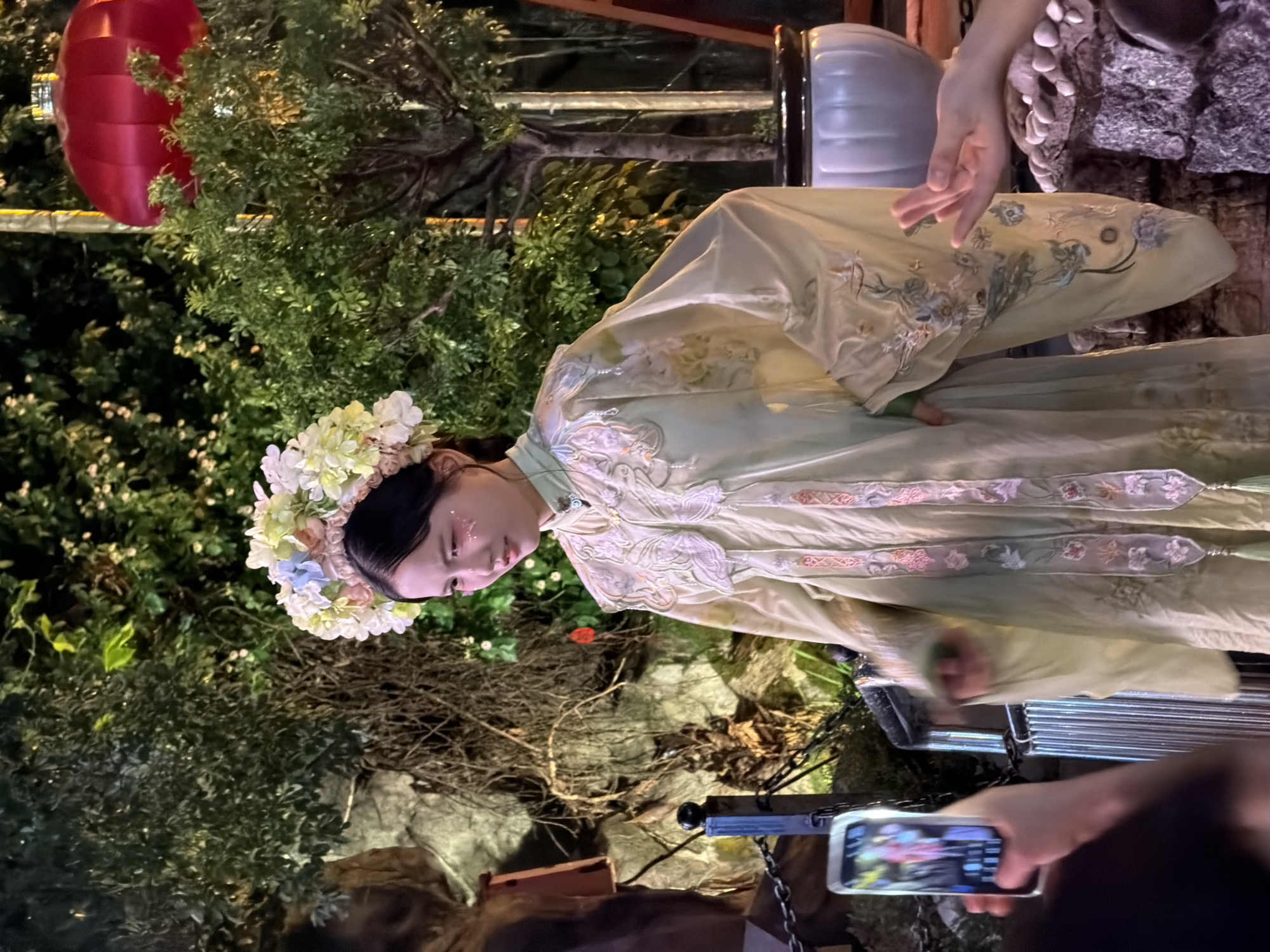 An East Asian woman in traditional Chinese attire, including a pale green Hanfu dress adorned with floral embroidery and a floral headdress, stands on a rocky, landscaped platform. Her facial expression is one of wistful contemplation as she looks slightly to her right. She has glitter makeup on her cheeks and near her eyes. In the background, lush green trees and foliage are visible, illuminated by soft lighting, suggesting an outdoor or garden setting at night. A large red lantern hangs from a pole on the right side of the frame. In the foreground, a person's hand holds a smartphone, capturing a photograph of the woman. Another hand, possibly belonging to a spectator, is visible near the bottom right corner of the image, reaching out towards the woman. The scene appears to be set in a public space, possibly a park or a cultural event, in the Yuzhong District of China.