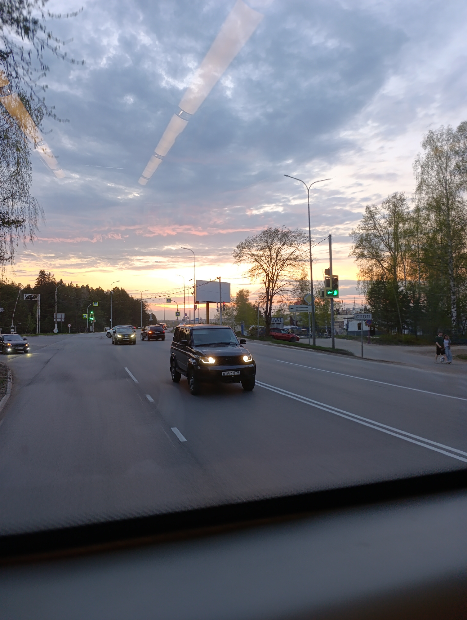 The image captures a street scene in Chaikovsky, Russia, at dusk. The sky displays a blend of grey clouds and orange hues from the setting sun. A black SUV is the primary subject, moving along a road with double white lane markers. Other vehicles are visible in the background, suggesting moderate traffic. Traffic lights show a green signal. Pedestrians are present on the right side of the road. Trees line the edges of the road, indicative of a residential or suburban area. A road sign and billboard are located in the middleground, contributing to the urban landscape. The image is taken from within a vehicle, with the dashboard visible at the bottom.