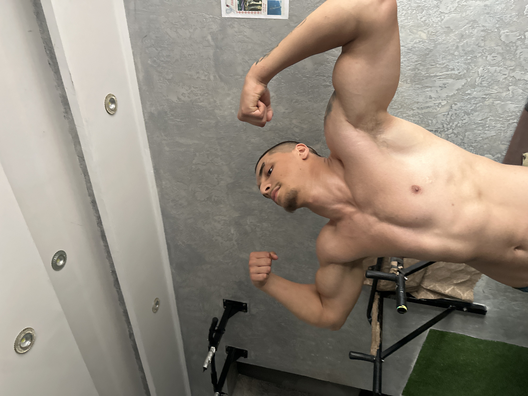 A muscular, shirtless man is posing in a gym, flexing his biceps. He has short, dark hair and a trimmed beard. The gym appears to have a pull-up bar, a gray textured wall, and a white wall with recessed lighting. A bulletin board with images is affixed to the gray wall. A section of artificial turf is visible on the floor near the pull-up bar.
