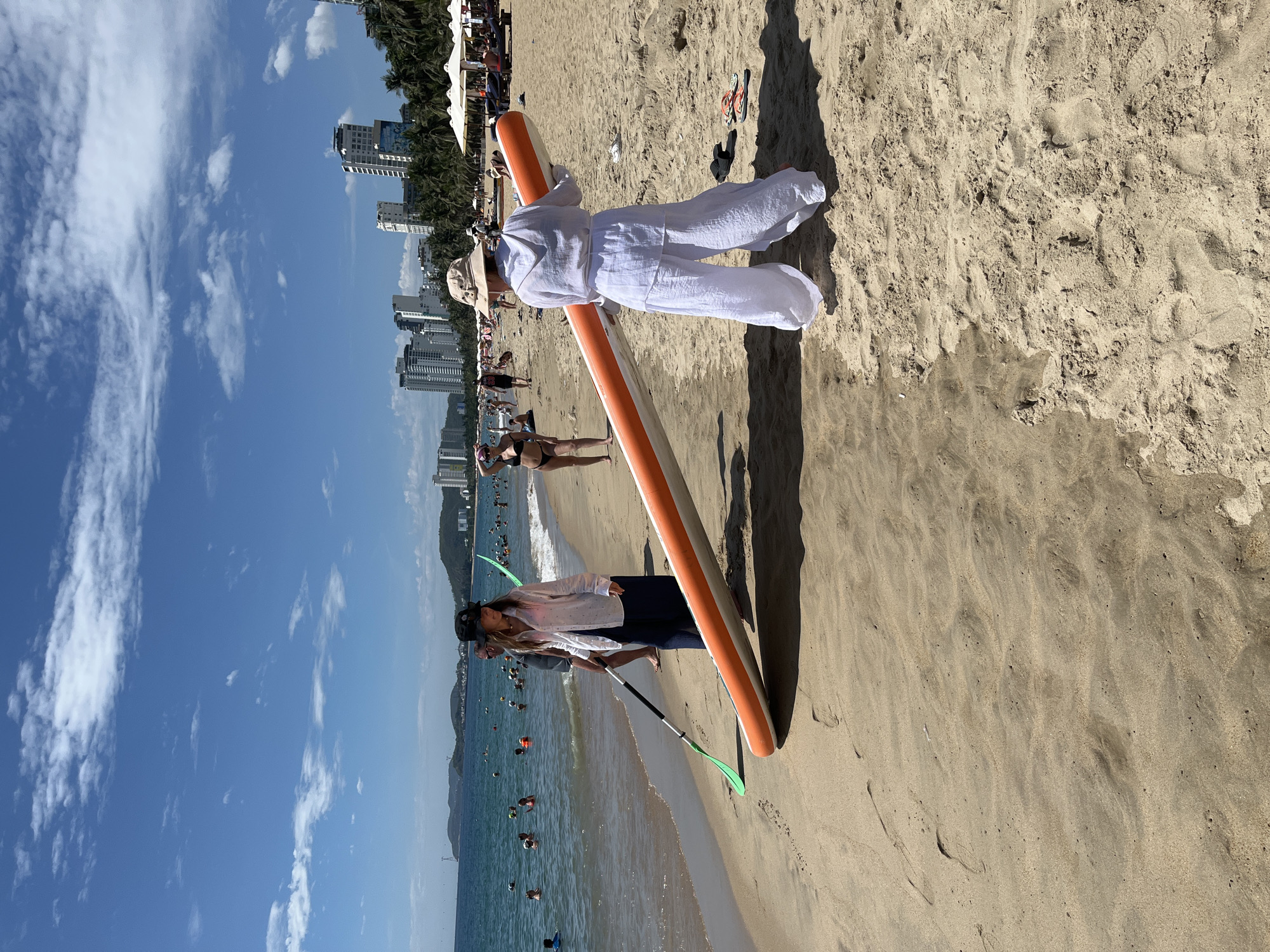 The image shows a sandy beach in Nha Trang, Vietnam, with the turquoise ocean in the background. The sky is blue with scattered white clouds. Several people are visible in the water and on the beach. In the foreground, two women are standing on the sand. One woman, wearing a white shirt and dark pants, holds a paddle. The other woman, dressed in white, carries a large orange and white paddleboard. In the background, tall buildings and palm trees line the coast. The water is dotted with swimmers and some small boats. The scene depicts a typical day at the beach, with people enjoying water activities and relaxing on the sand.