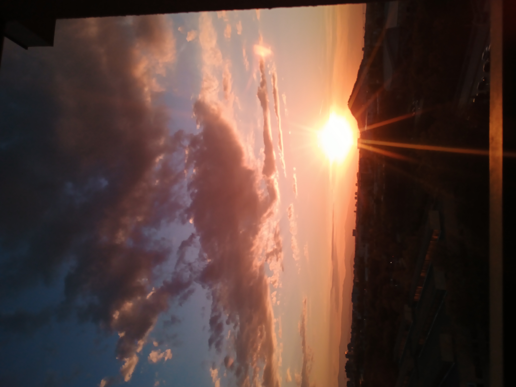 The image captures a sunset view from what appears to be a high vantage point, possibly a window or balcony, overlooking the city of Plovdiv, Bulgaria. The sky is filled with dramatic clouds illuminated by the setting sun, which dominates the scene with its bright orange glow. To the left, the sky transitions to a deeper blue with some clouds. The city skyline is visible as a dark, silhouetted mass in the distance. The photograph is taken from indoors, with parts of the frame or window visible at the edges. No people are visible in the photograph.
