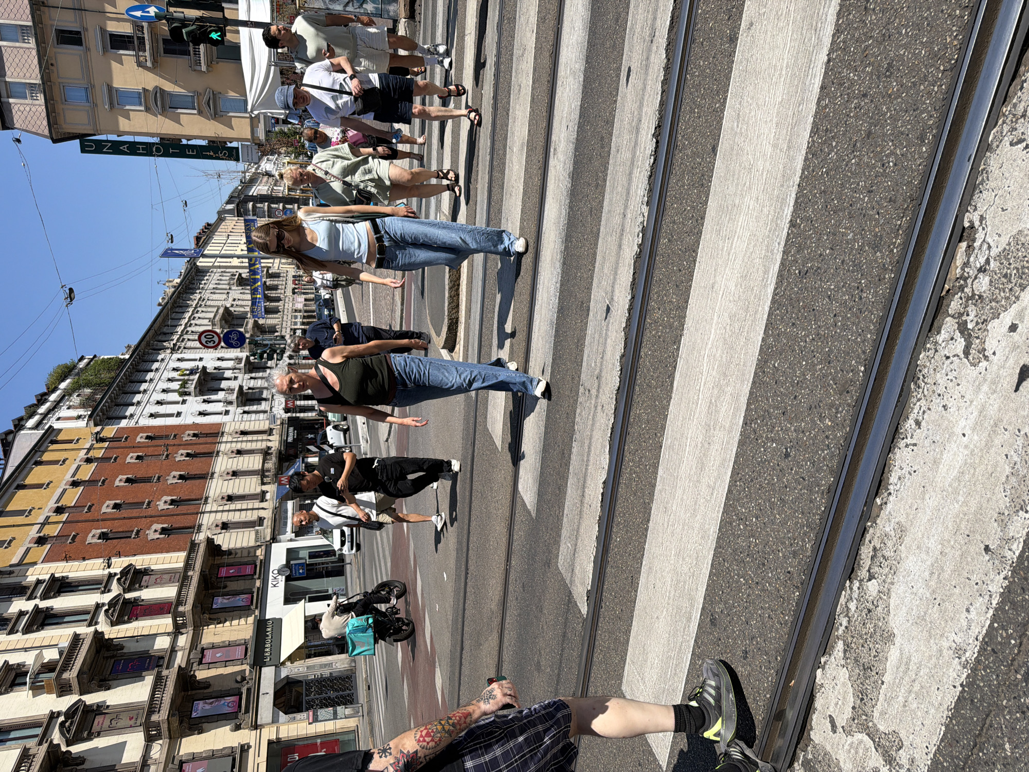 This is a street-level, medium shot of a crosswalk in Milan, Italy, on a bright, sunny day. The crosswalk is a busy pedestrian crossing with tram tracks running through it. Several pedestrians are crossing the street, including a group of young women in casual summer attire. One woman is wearing a light blue tank top and jeans, another a green tank top and jeans, and a third a light green shirt and shorts. They appear to be walking together. In the background, the architecture of Milan is visible, with multi-story buildings lining the street. Some of these buildings feature traditional European architectural elements, such as balconies and decorative facades. Cars and trams are present on the street, indicating a typical urban environment. The scene captures a moment of daily life in the city.