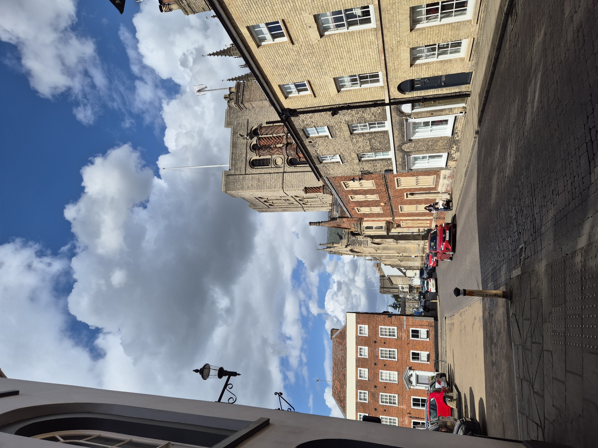 The image captures a street scene in West Suffolk, United Kingdom. The view is oriented along a street lined with multi-story brick buildings, some of which are light beige or yellowish in color. These buildings feature numerous windows with white frames. The tower of a church is visible between the buildings. The street is paved with cobblestones. Several cars are parked along the side of the street. The sky above is partly cloudy. A lamp post is located on the lower left corner. A yellow pole sits at the edge of the road. Two people walk by in the middle distance.