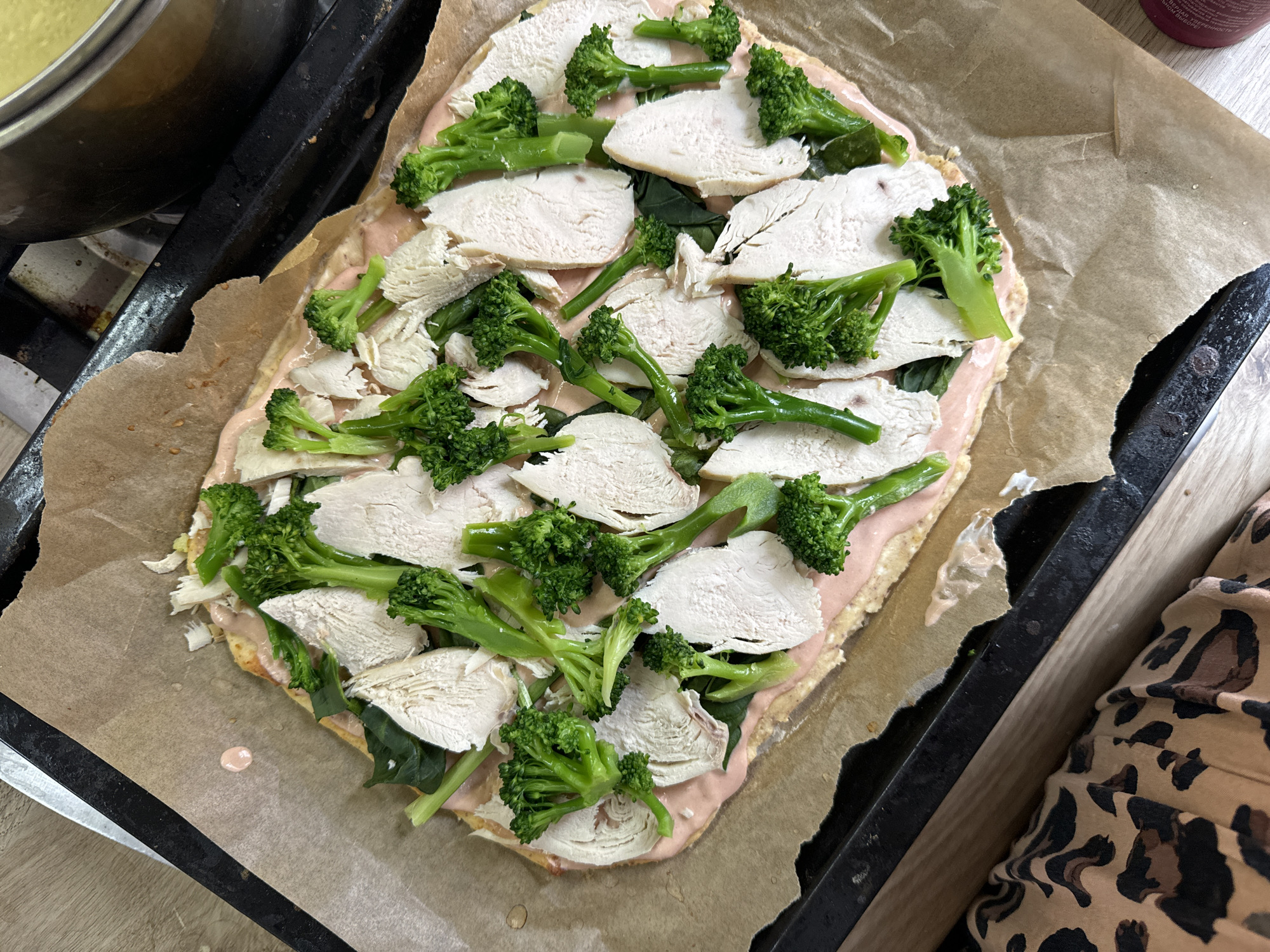 The image shows an uncooked rectangular pizza on parchment paper, placed on a black baking sheet. The pizza is topped with a pink sauce, spinach leaves, sliced chicken, and broccoli florets. A stainless-steel pot is visible in the upper left corner. A portion of a person's garment with a leopard print is present in the lower right corner. The image provides no information to determine its location.