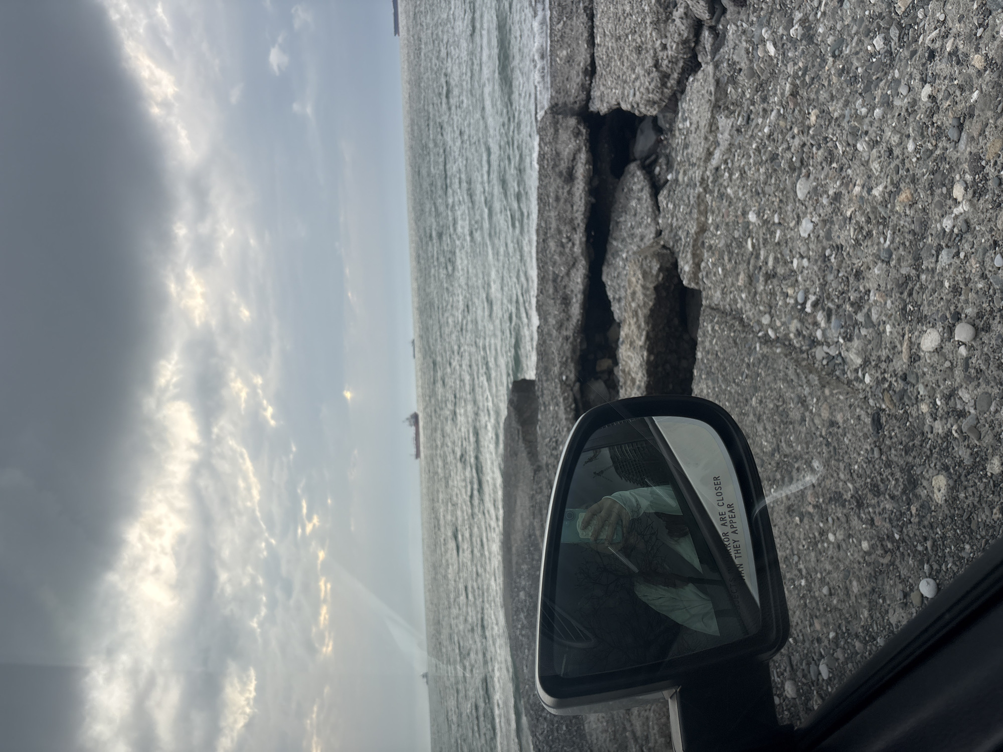 The photograph, taken from inside a car, looks out towards the sea. A car's side mirror is visible, reflecting the person holding a cell phone taking the picture. The sea appears grey and textured. The sky above is overcast with dark clouds on the left. A cracked, stone barrier divides the view from the sea. The location is likely in Batumi, Georgia, given the coastal environment. Several ships are visible on the horizon, suggesting a busy maritime environment.