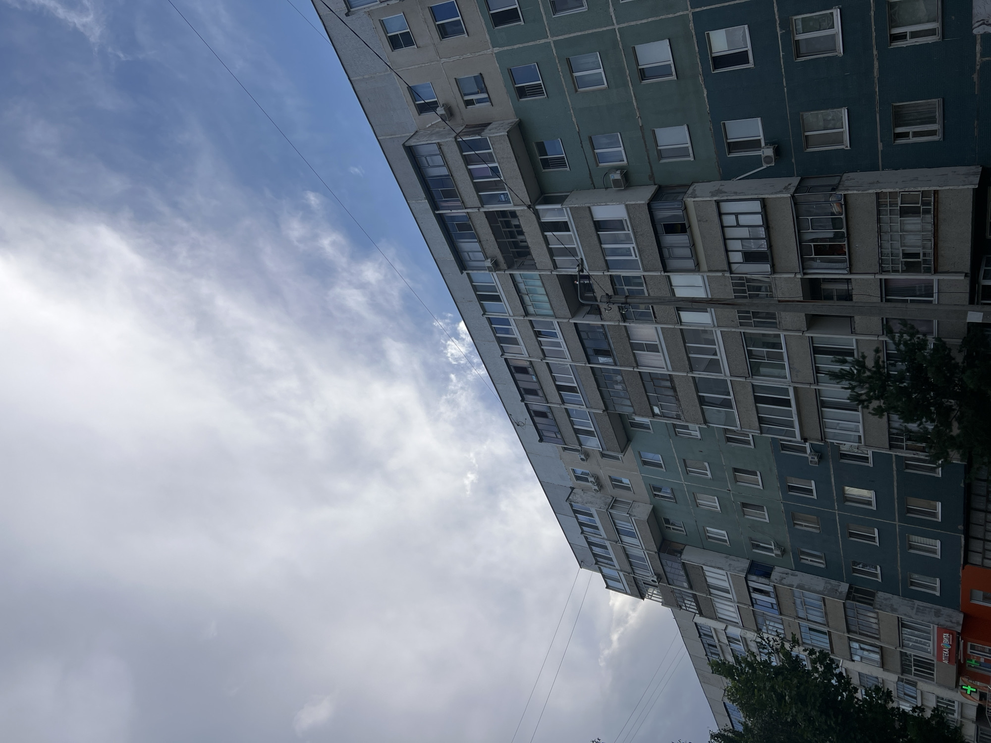 The image depicts a multi-story apartment building under a partly cloudy sky. The building features a mix of blue-green and grey facades, with numerous windows of varying sizes and configurations, including balconies with glass enclosures. Several air conditioning units are visible on the exterior walls. A utility pole with wires runs vertically in front of the building. In the lower left corner, a sliver of green foliage from a tree is visible, and a storefront with a red awning and signage reading "АПТЕКА ВИТА" (Pharmacy Vita) is partially visible. The scene appears to be an urban residential area. Without additional information or context, the precise location within Ulyanovsk, Russia, cannot be determined from the image alone.