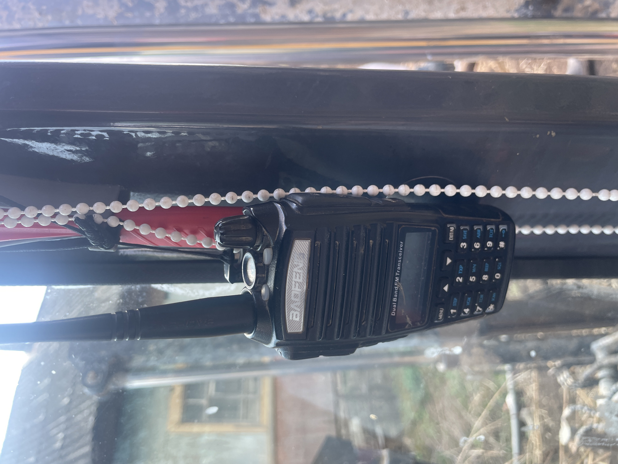 A Baofeng Dual Band FM Transceiver is mounted inside a vehicle, likely an excavator or heavy machinery due to the rugged environment visible through the windshield. The radio is positioned near a white beaded chain. Outside, the environment suggests a rural or industrial location in the city of Поедуги, Russia. Visible through the glass is an indistinct building, and the surroundings appear to be slightly unkempt with what seems to be foliage or brush.