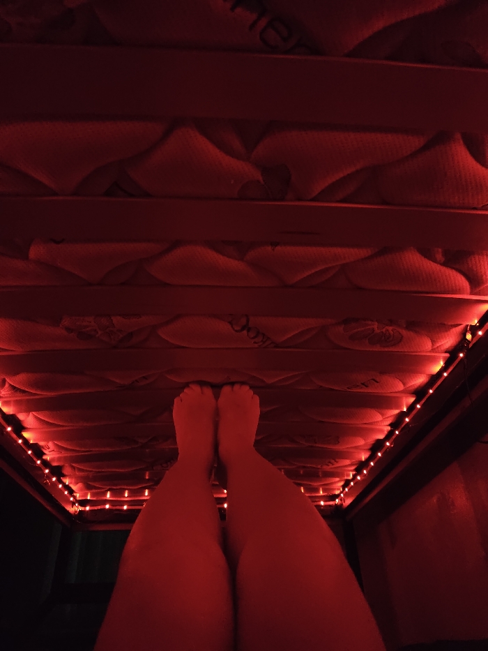 A pair of human legs and feet are visible, illuminated by red light, positioned as if lying or sitting beneath a raised structure. Above the legs, the underside of a mattress with visible quilting patterns and branding is present. Horizontal bed slats support the mattress. Along the perimeter of the bed frame, a string of red LED lights is installed, casting a strong red hue across the entire scene. The environment is dark, with the primary illumination originating from these red LED strips. The location in the city of Chaikovsky, Russia, cannot be determined from the image provided.
