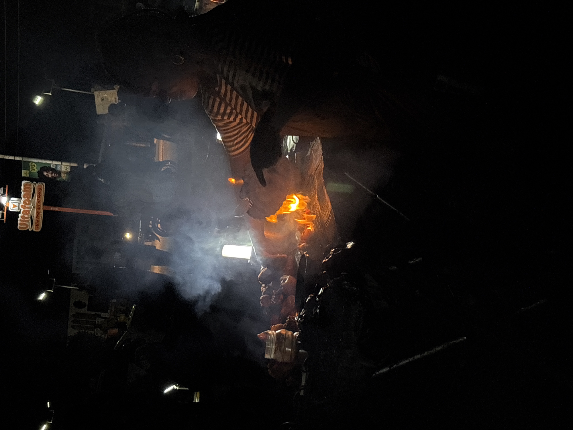A person wearing a striped shirt tends to a vertical spit of meat, the flames rising and smoke billowing around them. The scene appears to be at night in a city, possibly Barakin Lerawi, Nigeria, indicated by a sign in the background. The surrounding environment is dark.