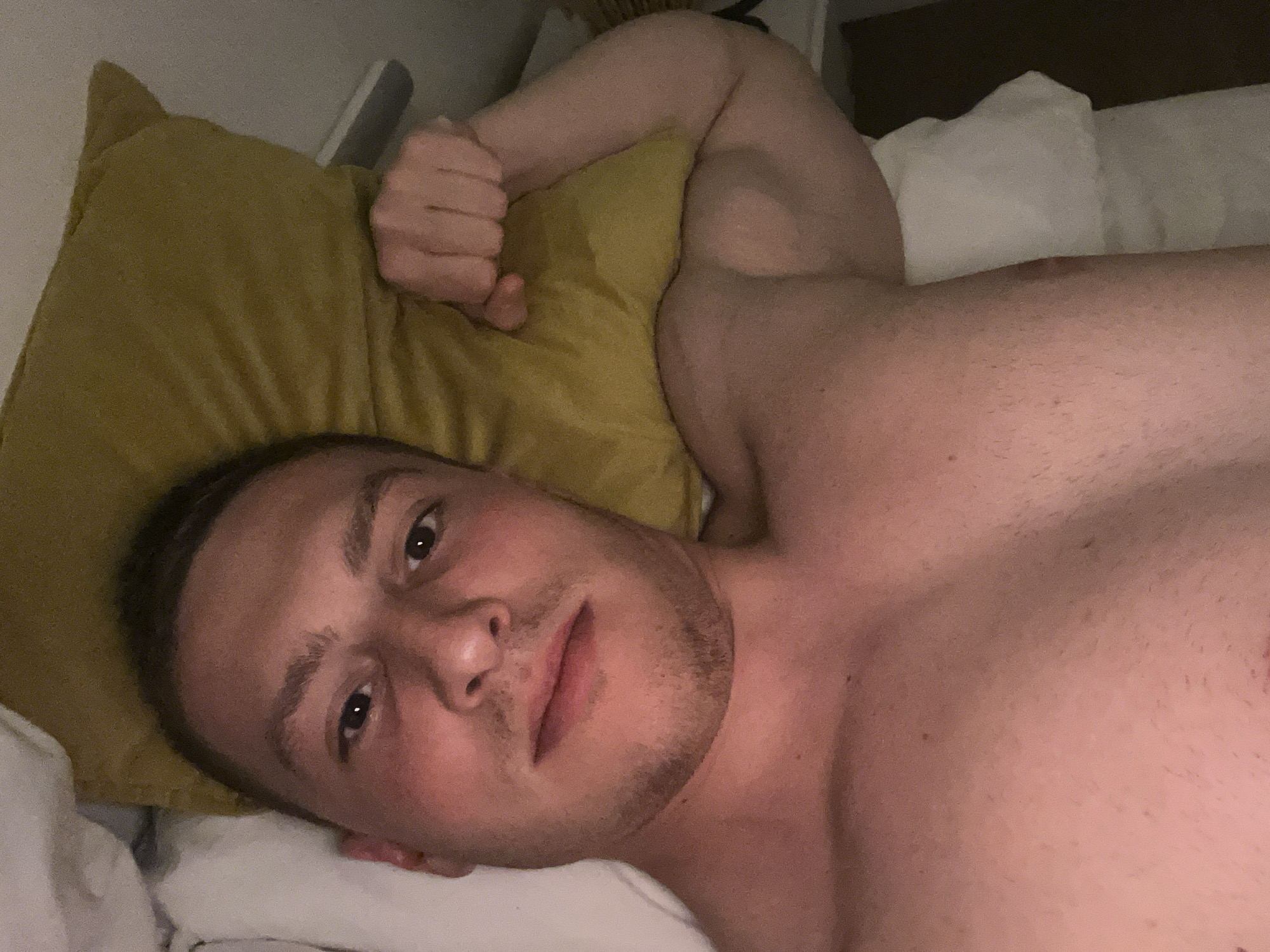 A young man is depicted lying down, shirtless, in what appears to be a bed. He is positioned on his back, looking directly towards the camera with a neutral and calm expression. His head rests on a white pillow, with a mustard-yellow pillow placed next to it. His right arm is bent at the elbow, with his fist lightly clenched, resting on the yellow pillow. The man has short, dark hair and visible stubble on his chin and cheeks. The lighting in the image is dim, suggesting an indoor setting like a bedroom, possibly during the evening or night. White bedding and additional pillows are visible around him. His bare torso and shoulders are visible. No specific location cues are present within the image itself, but the provided context is Vitry-sur-Seine, France. There is no visible text in the media file.