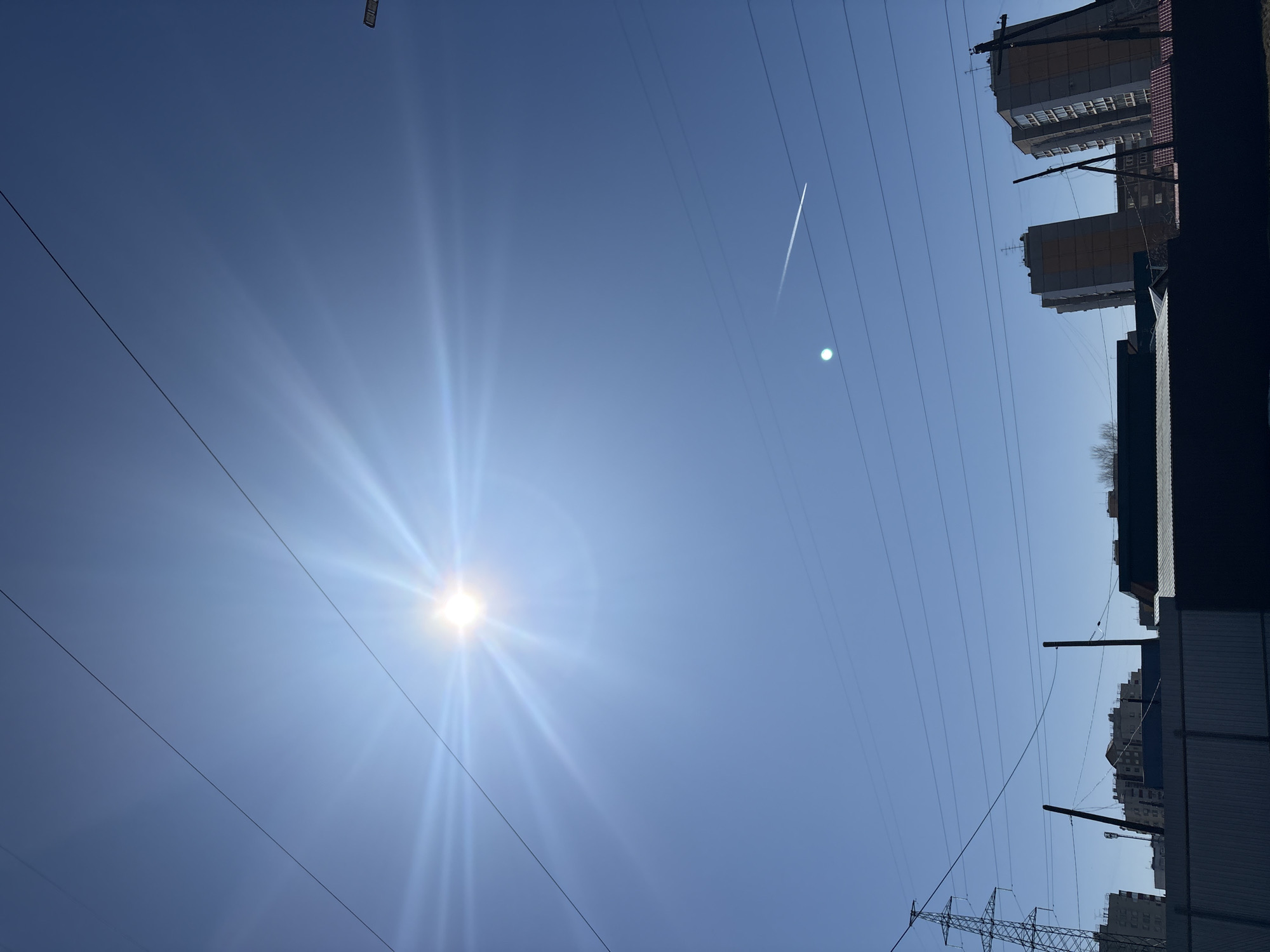 The image shows a bright, blue sky with the sun visible as a point source with radiating light rays. There are several parallel electrical wires stretching across the sky from lower left to upper right. A white contrail is visible diagonally across the upper portion of the sky. Buildings are visible along the top and right edges of the frame, partly obscured by shadows. An electrical tower is in the lower right corner. This image was taken in Chaikovsky, Russia.