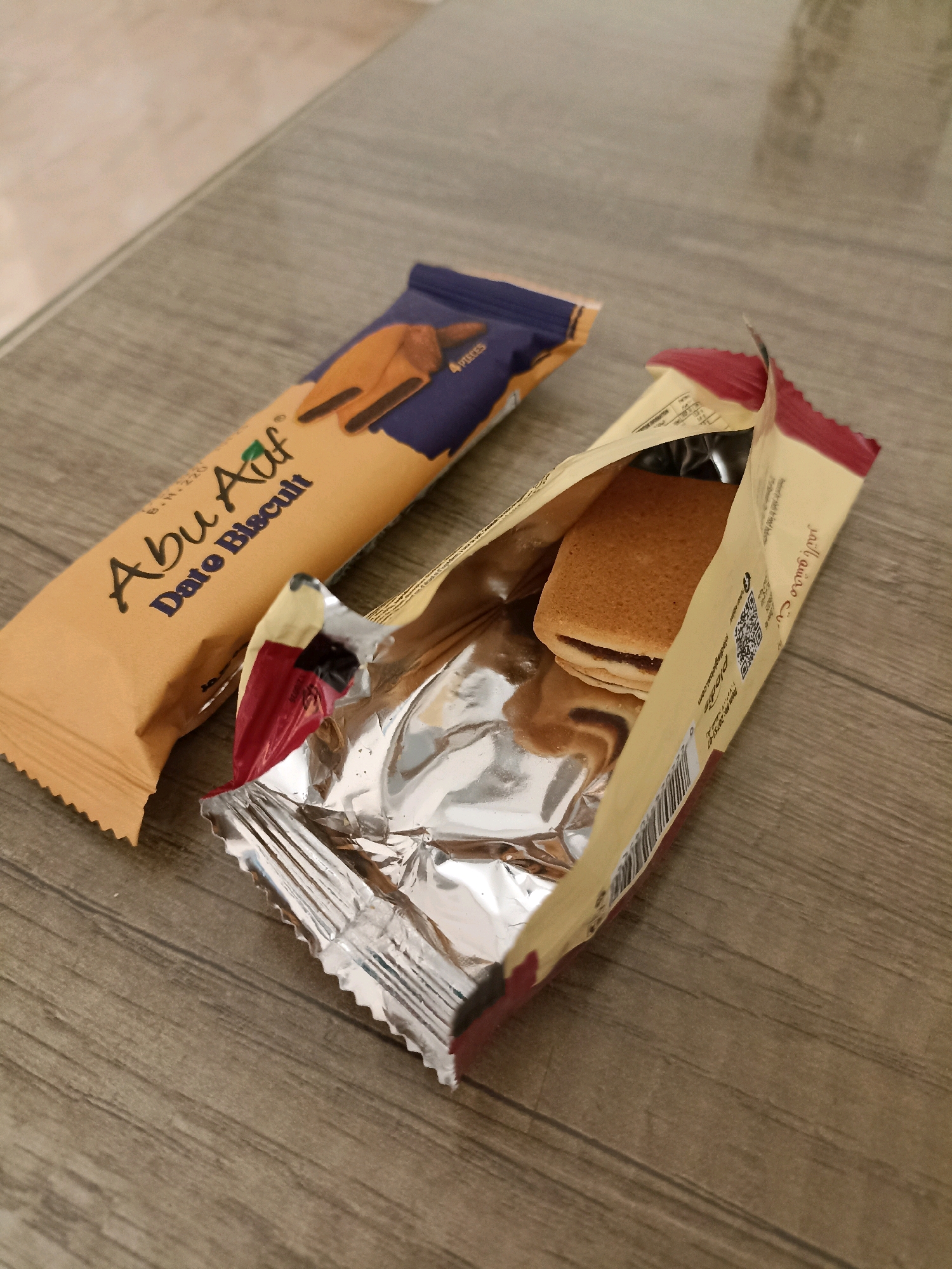 Two packages of Abu Auf Date Biscuits are on a wooden table. One package is sealed and lies to the left, featuring a brown and blue wrapper with the brand name and product type printed in Arabic and English. The other package is open and lies to the right, revealing two biscuits inside. The biscuits are rectangular with a filling. The scene appears to be indoors, possibly in Al Hadaba, Egypt, given the context. The lighting suggests natural light coming from an unseen source.