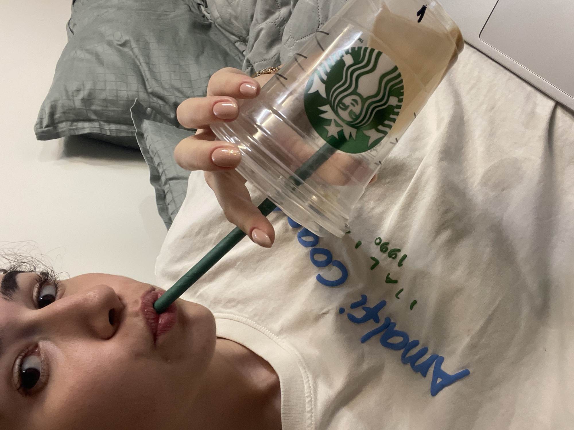 A young woman lies on her back, holding a Starbucks cup with a green straw to her lips. The cup is mostly empty, with a small amount of liquid at the bottom. She is wearing a white t-shirt with the word "Amalfi" written in blue and the number "1990" written in green. A gray pillow is visible in the background. A laptop rests on the fabric beside the cup. There is no indication of the location being Sofia, Bulgaria.