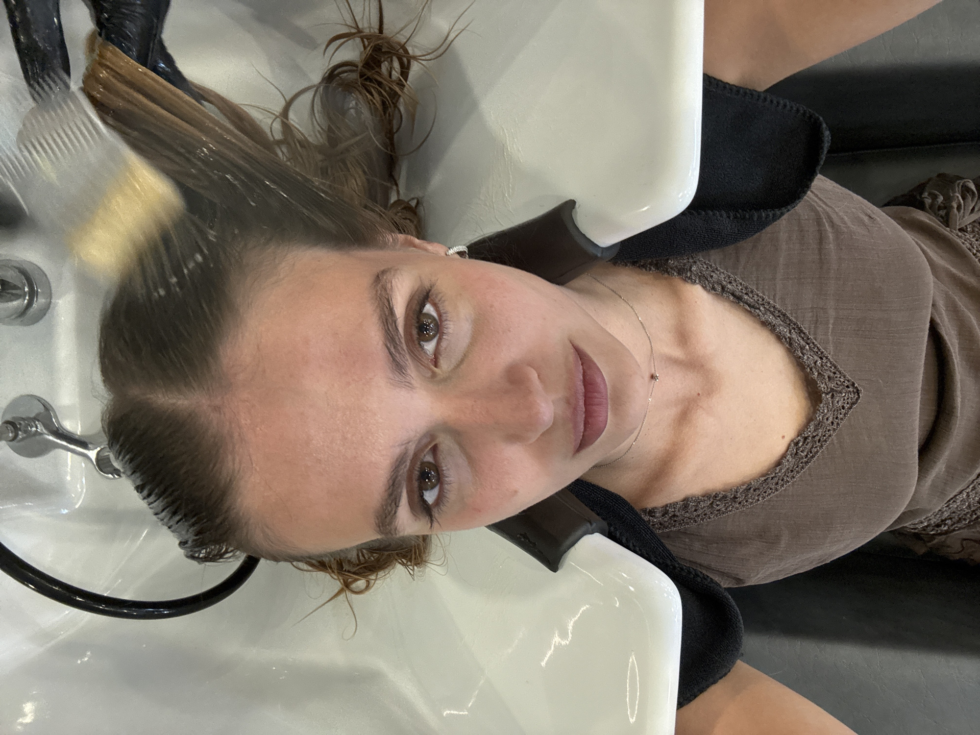 A woman is at a hair salon in Tbilisi, Georgia. She is reclining in a hair washing station. Her wet hair is being rinsed in a white porcelain sink. She is wearing a brown top with a lace neckline. A black fabric neck guard is around her neck. A silver faucet is visible to the left. She has fair skin, brown eyes, and is wearing a silver necklace and earring.